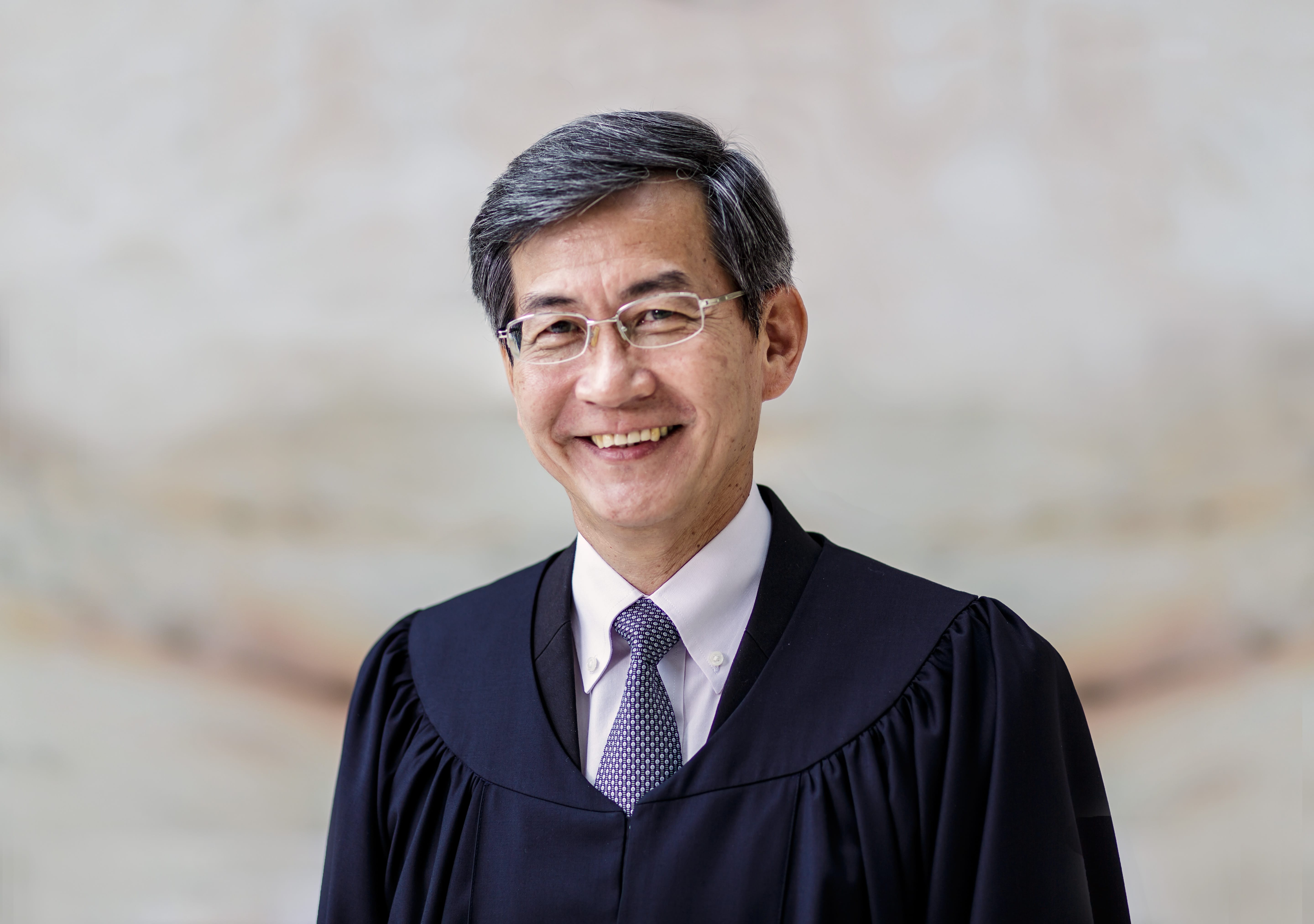 Man in judicial robe, glasses, smiling. White shirt, patterned tie.