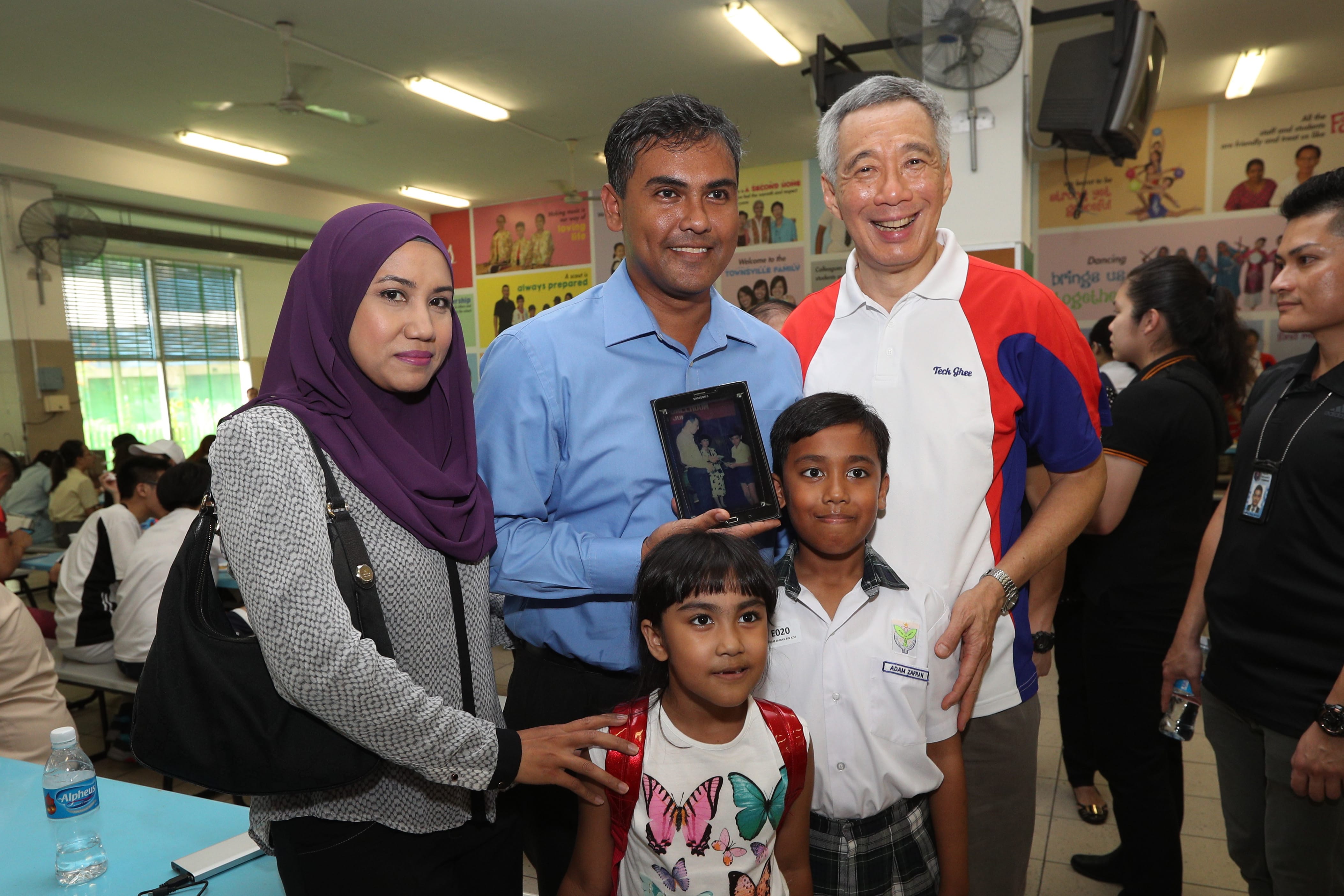 Group poses indoors. Man holds a Samsung tablet showing a photo. Lee Hsien Loong is present.