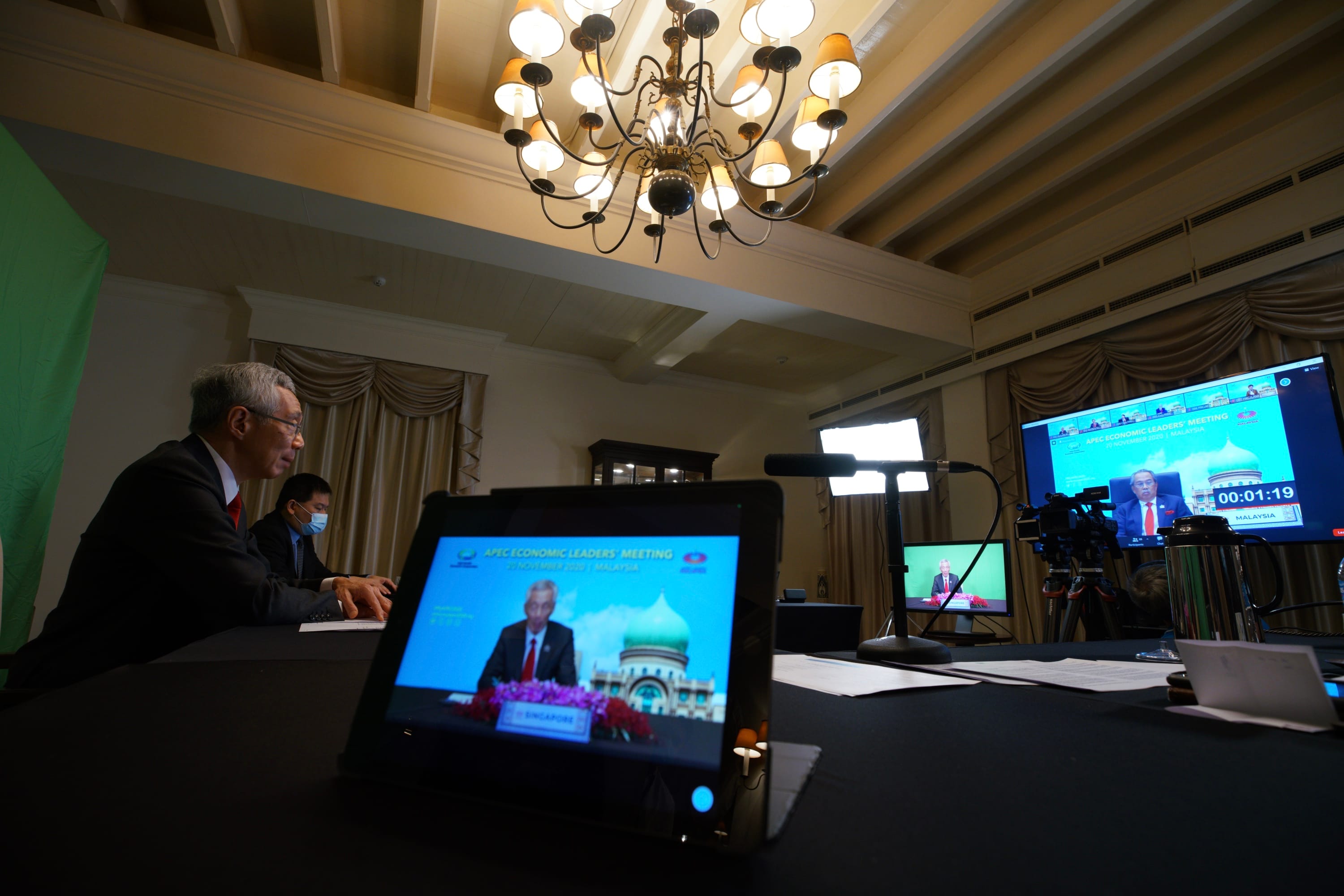 Conference room with men, screens showing APEC Leaders' Meeting, and recording equipment.