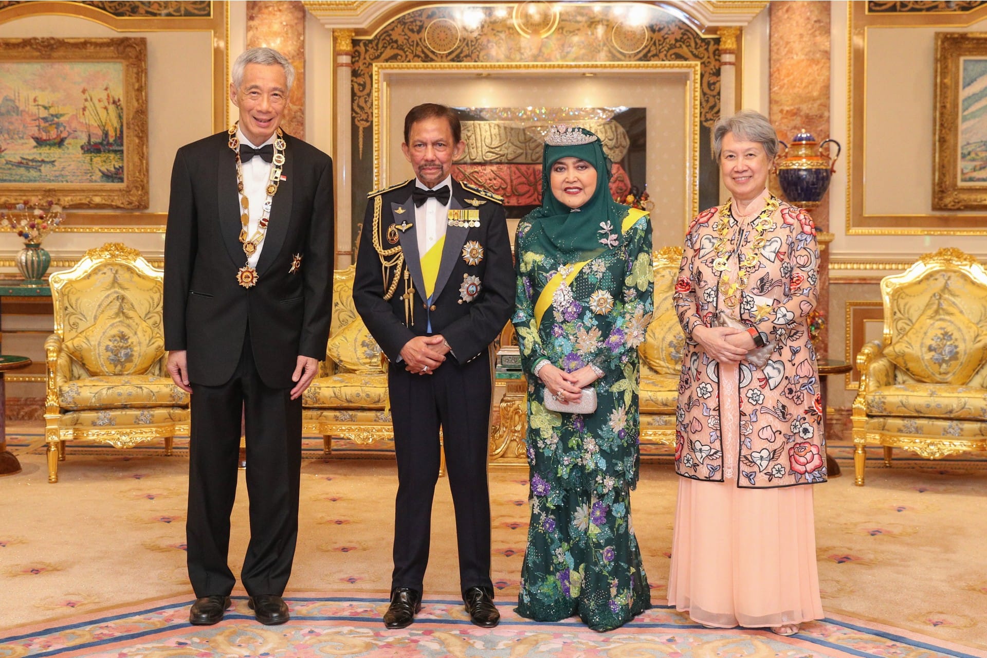 Four people stand in an ornate room with gold furnishings and decorative carpet.