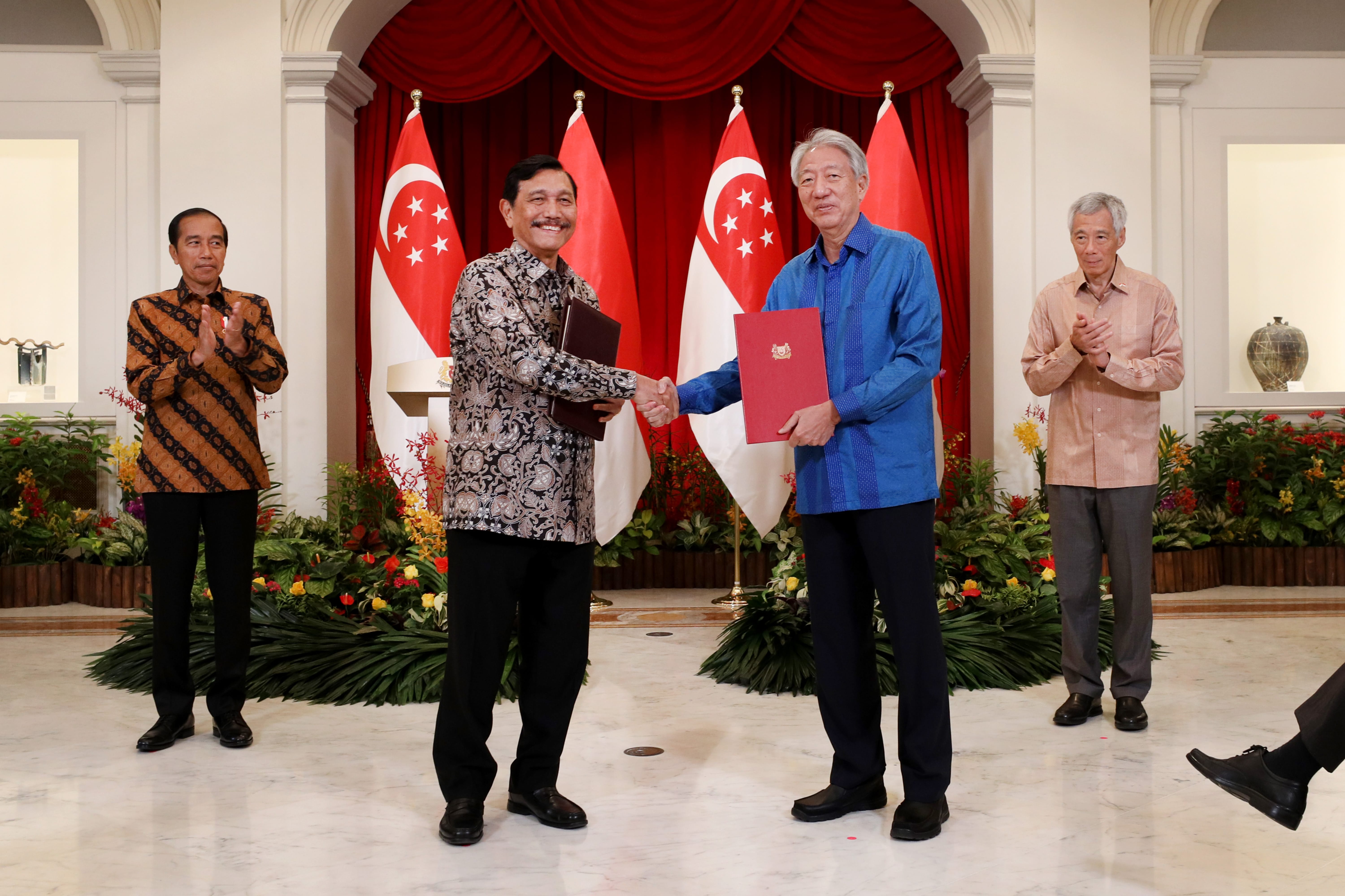 Two men shake hands; two others stand nearby, clapping, before Singapore flags.