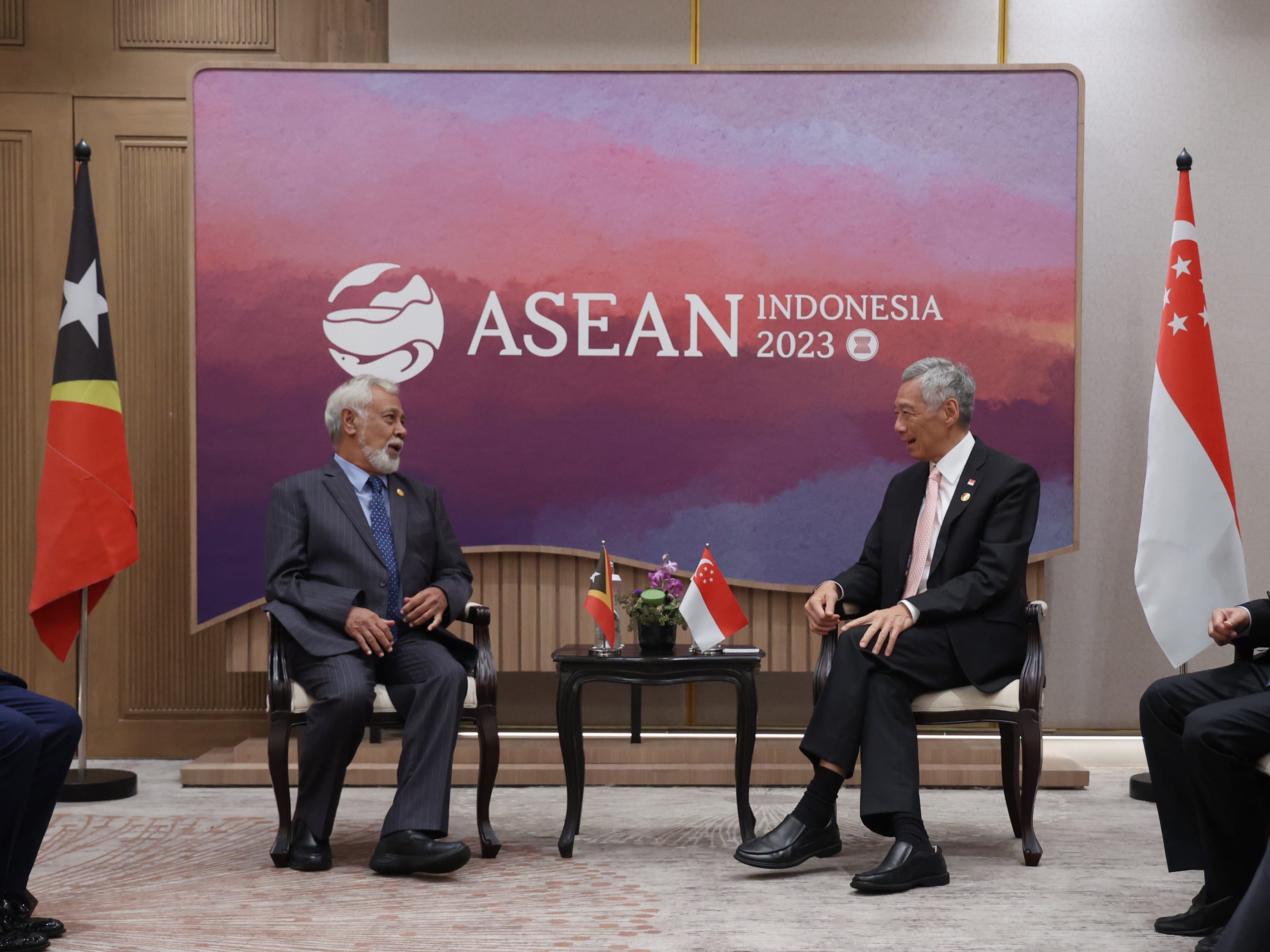 Two men in suits seated, facing each other with ASEAN Indonesia 2023 backdrop and Timor-Leste, Singapore flags.