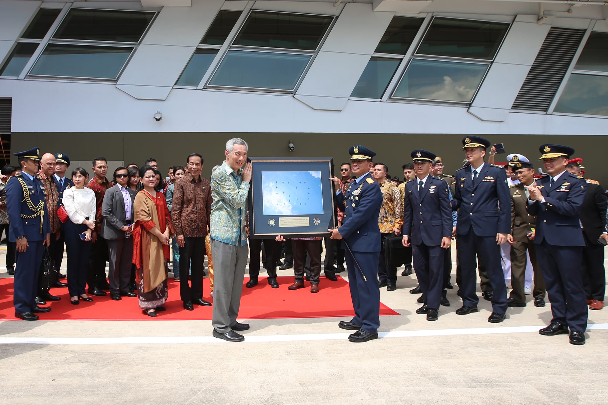 Lee Hsien Loong receives framed aircraft photo from military officer, crowd and building behind.