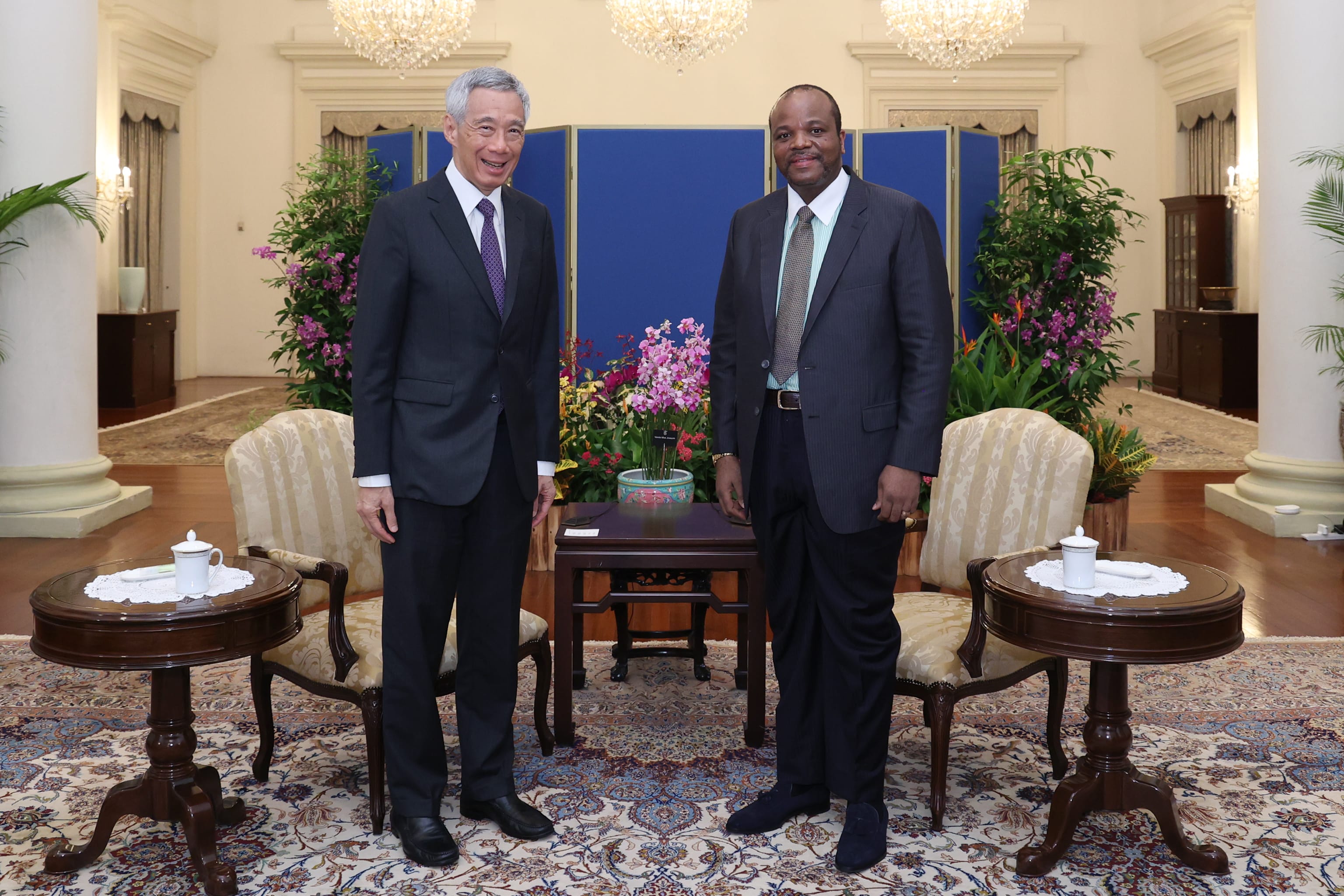 Two men in suits stand side-by-side indoors amid floral arrangements and furniture.