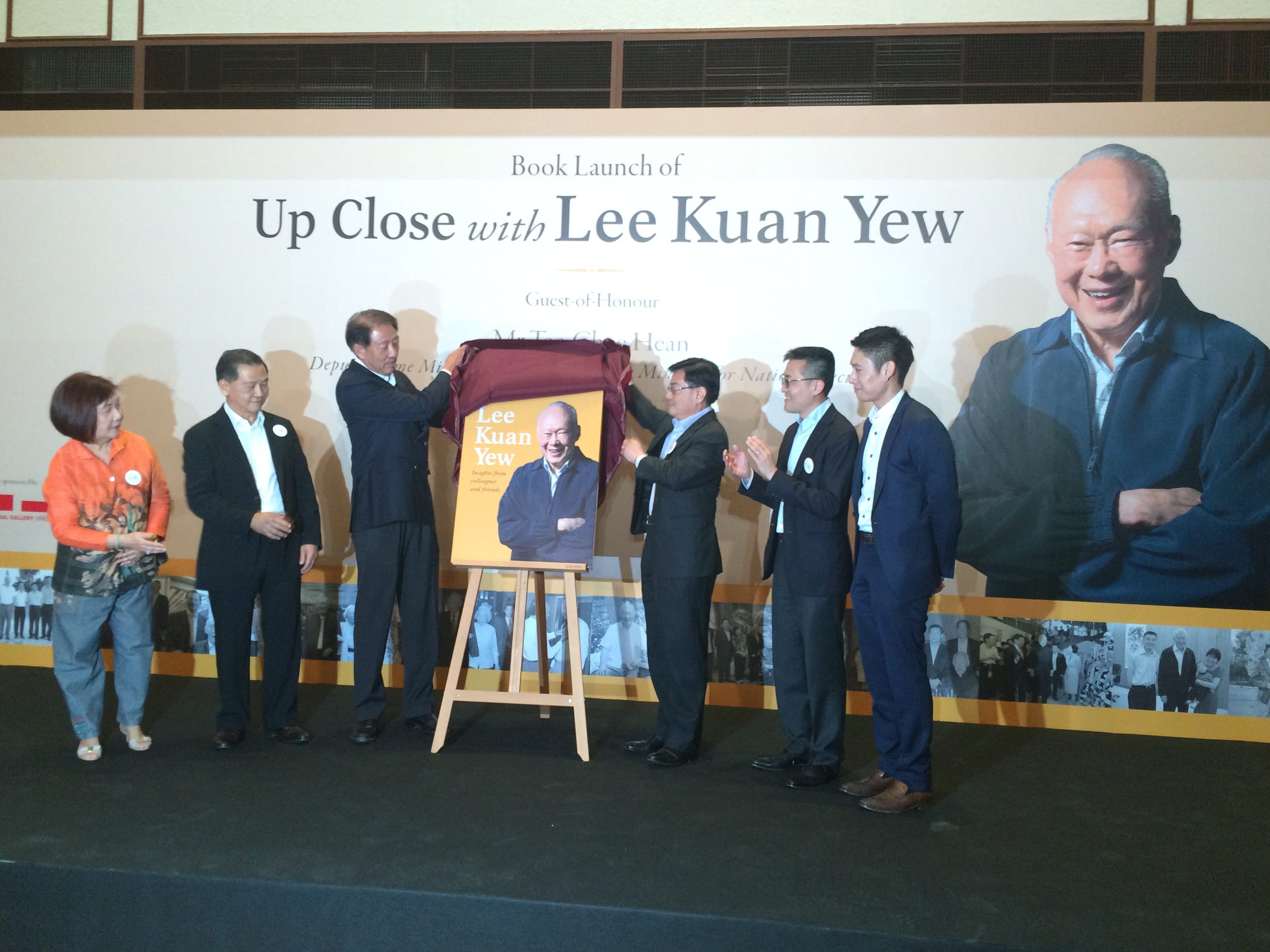 Book launch event: People unveil "Up Close with Lee Kuan Yew" book cover on easel. A large Lee Kuan Yew image behind.