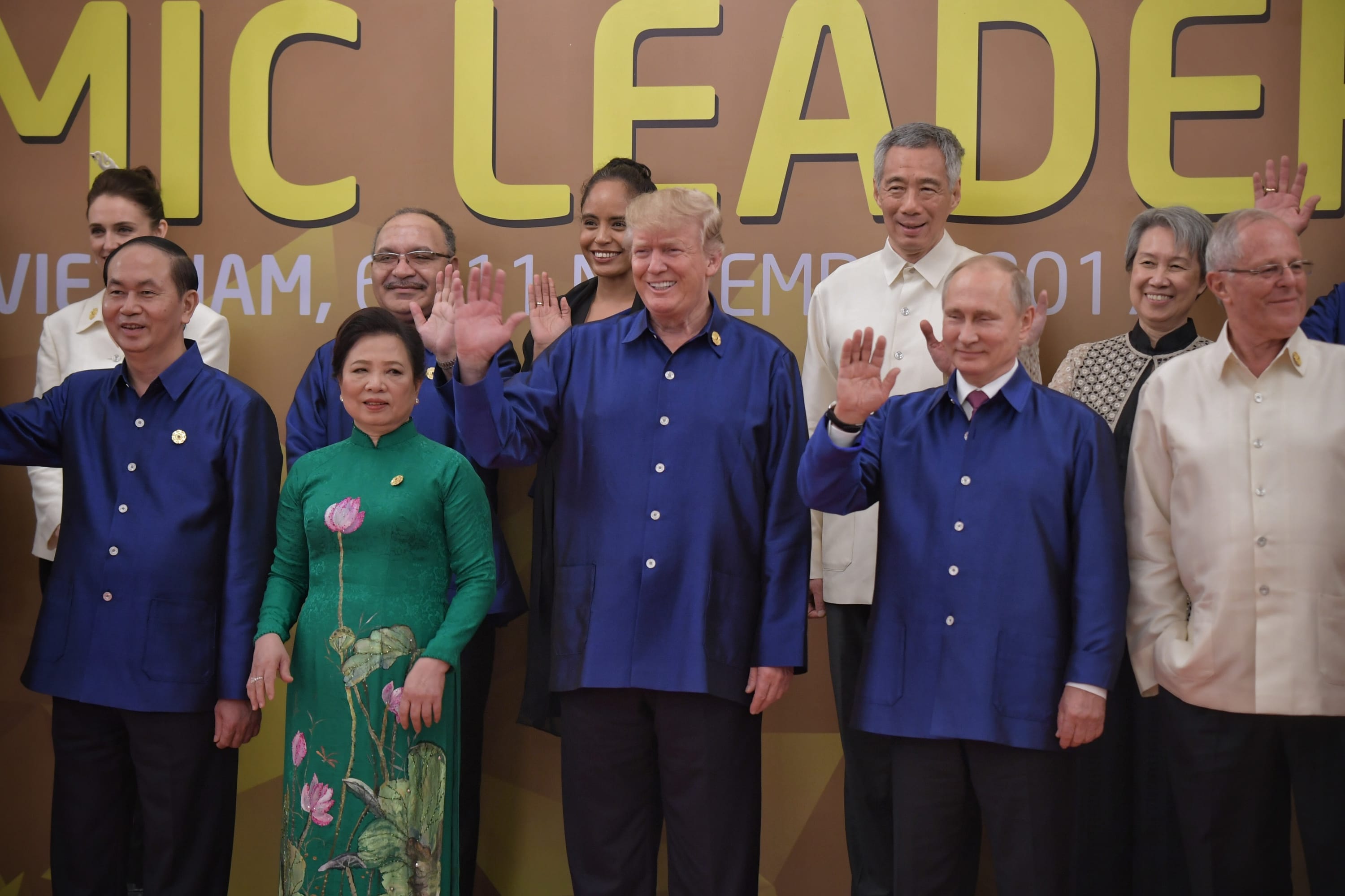 Group of people, including Trump and Putin, waving in front of a "MIC LEADER" backdrop.