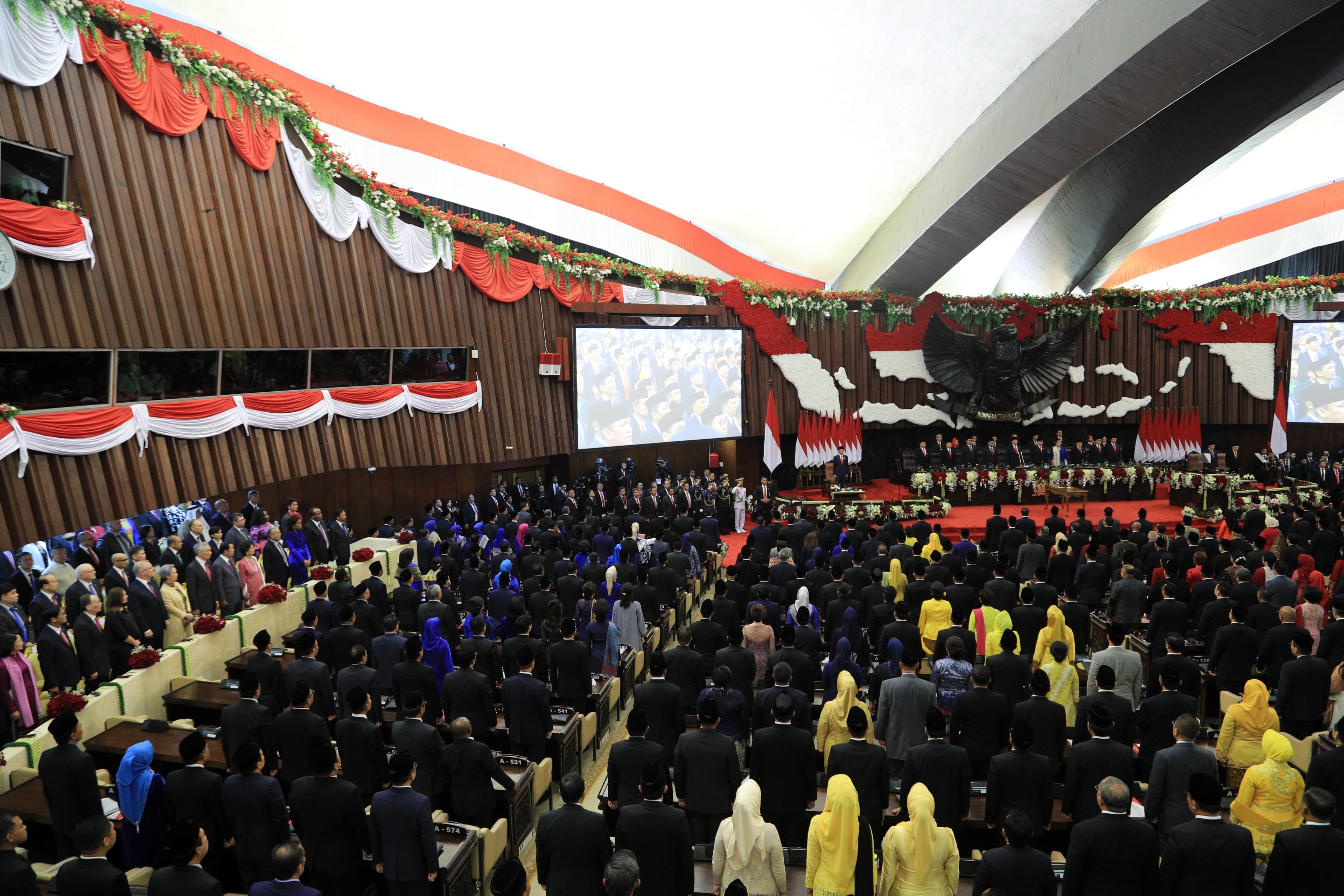 Large audience in a decorated hall. Red/white banners, screens, and Garuda Pancasila.