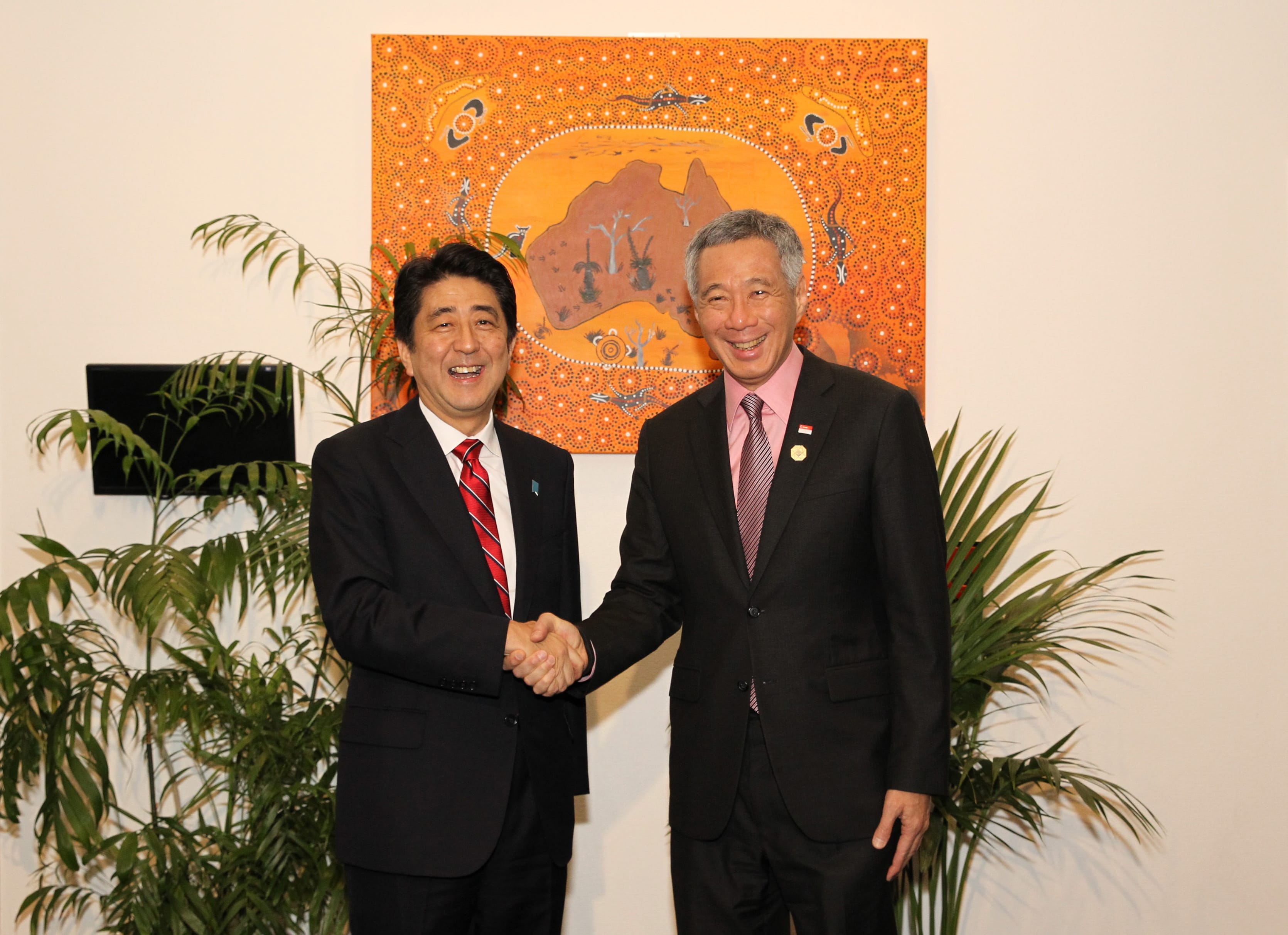 Shinzo Abe & Lee Hsien Loong shaking hands, wearing suits, in front of an orange Australian aboriginal-style painting.