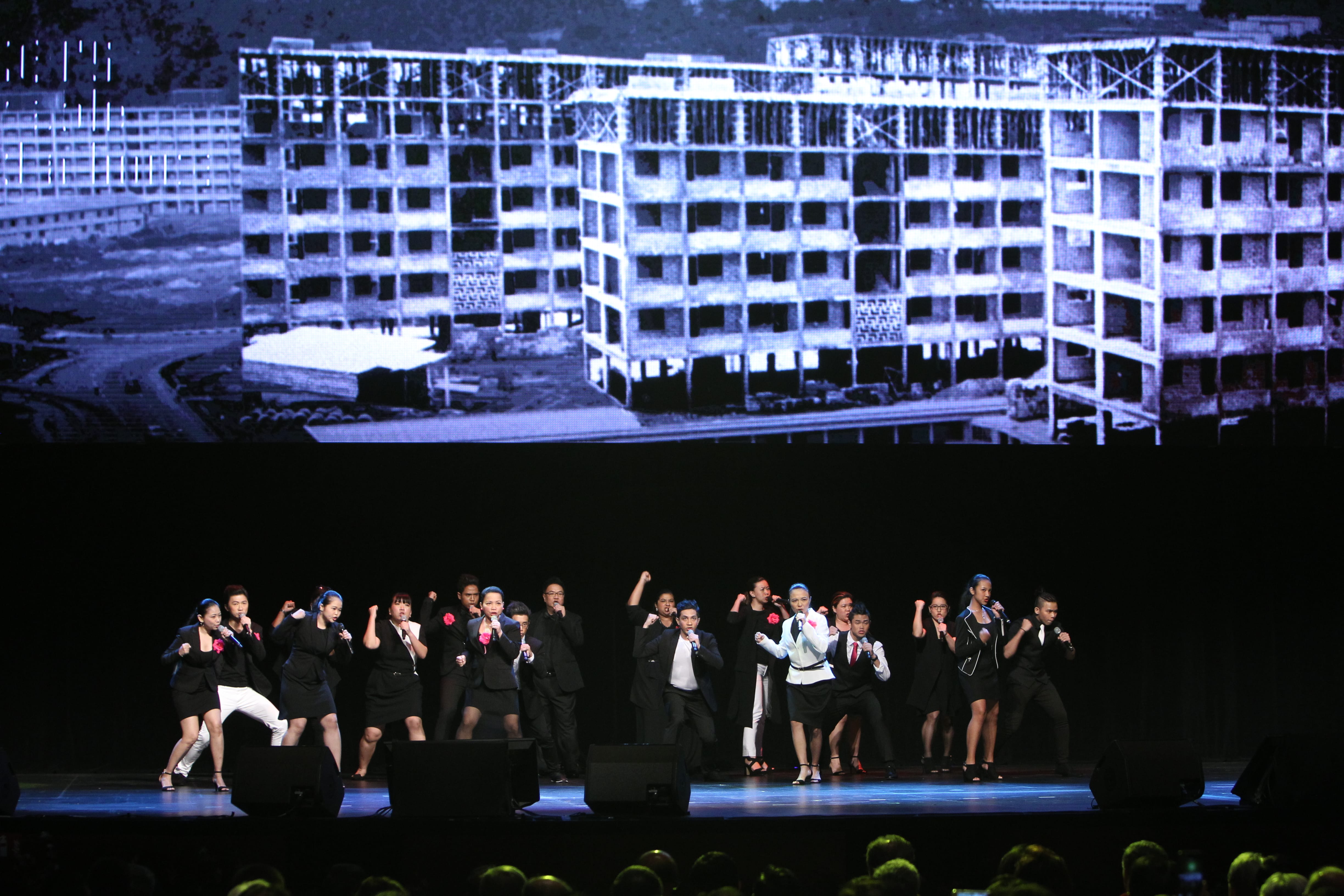 Group in suits performs on stage with mics, building construction video backdrop.