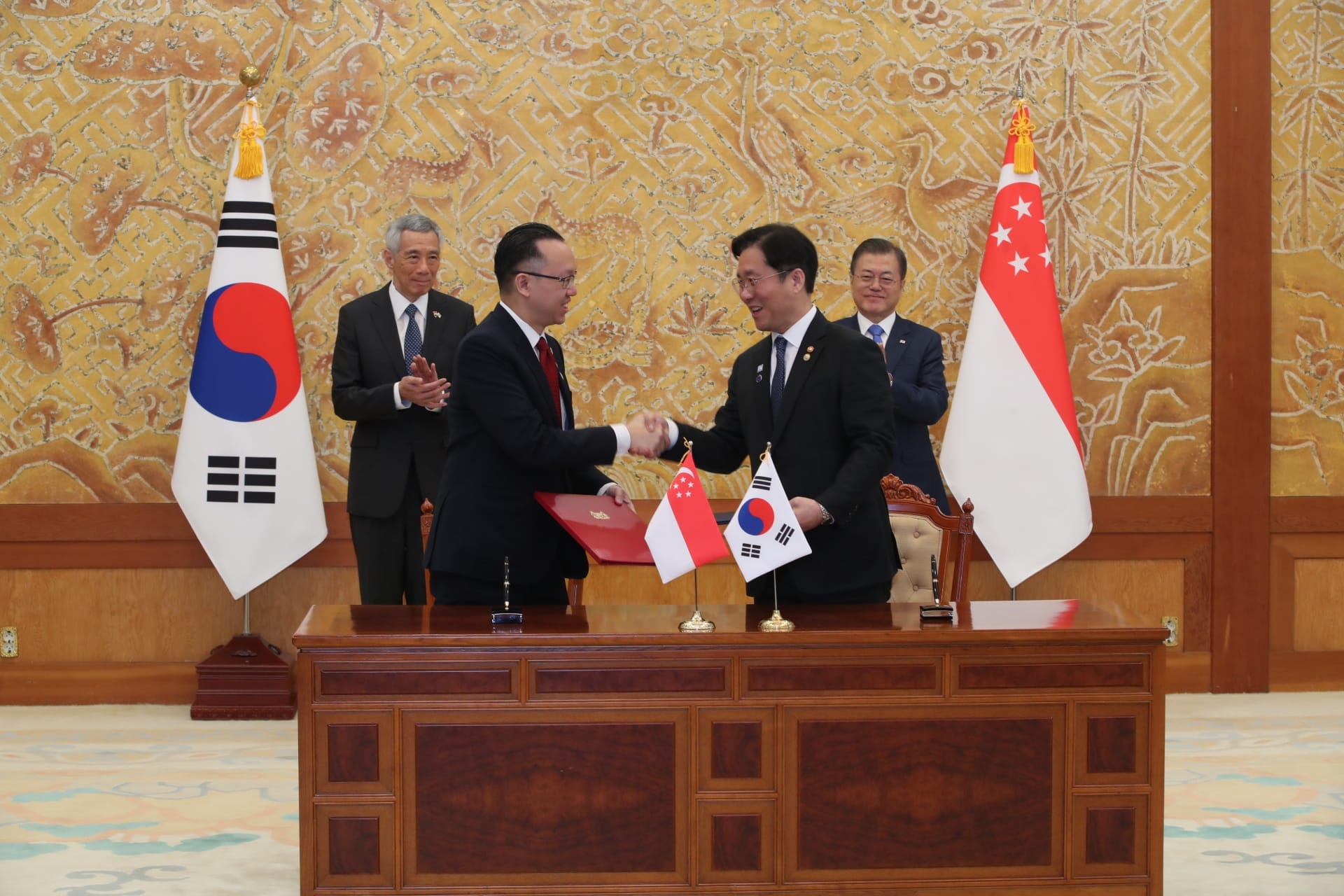 Two men in suits shake hands with flags of Korea and Singapore on a wood table.