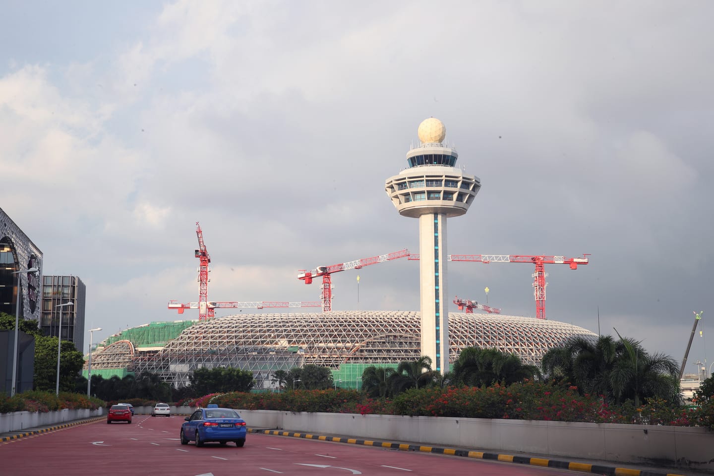 Airport control tower and dome structure under construction, with red cranes, above a red road with cars.