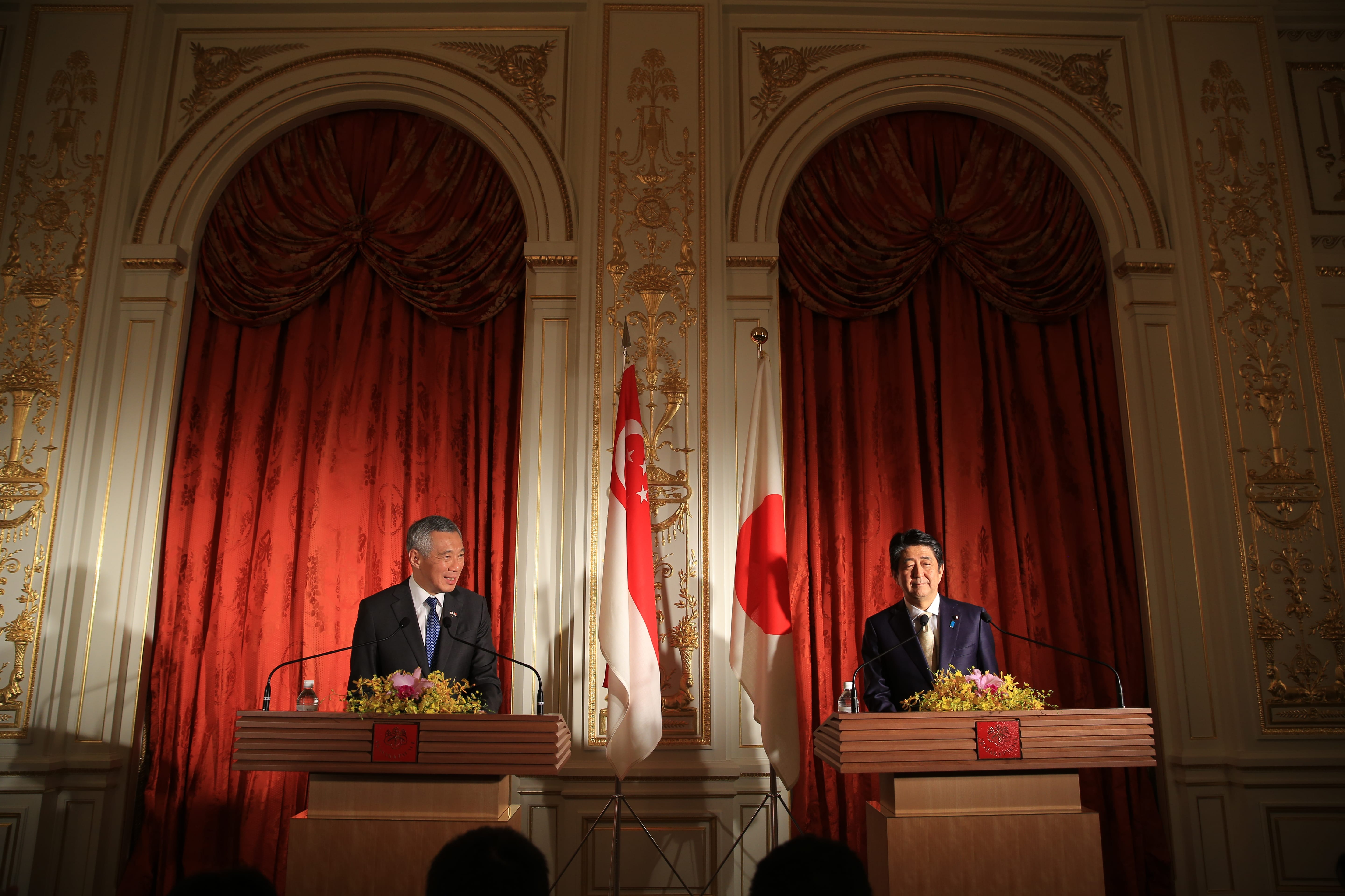 Two men stand at podiums with Singapore and Japanese flags. Ornate gold walls surround red curtains.
