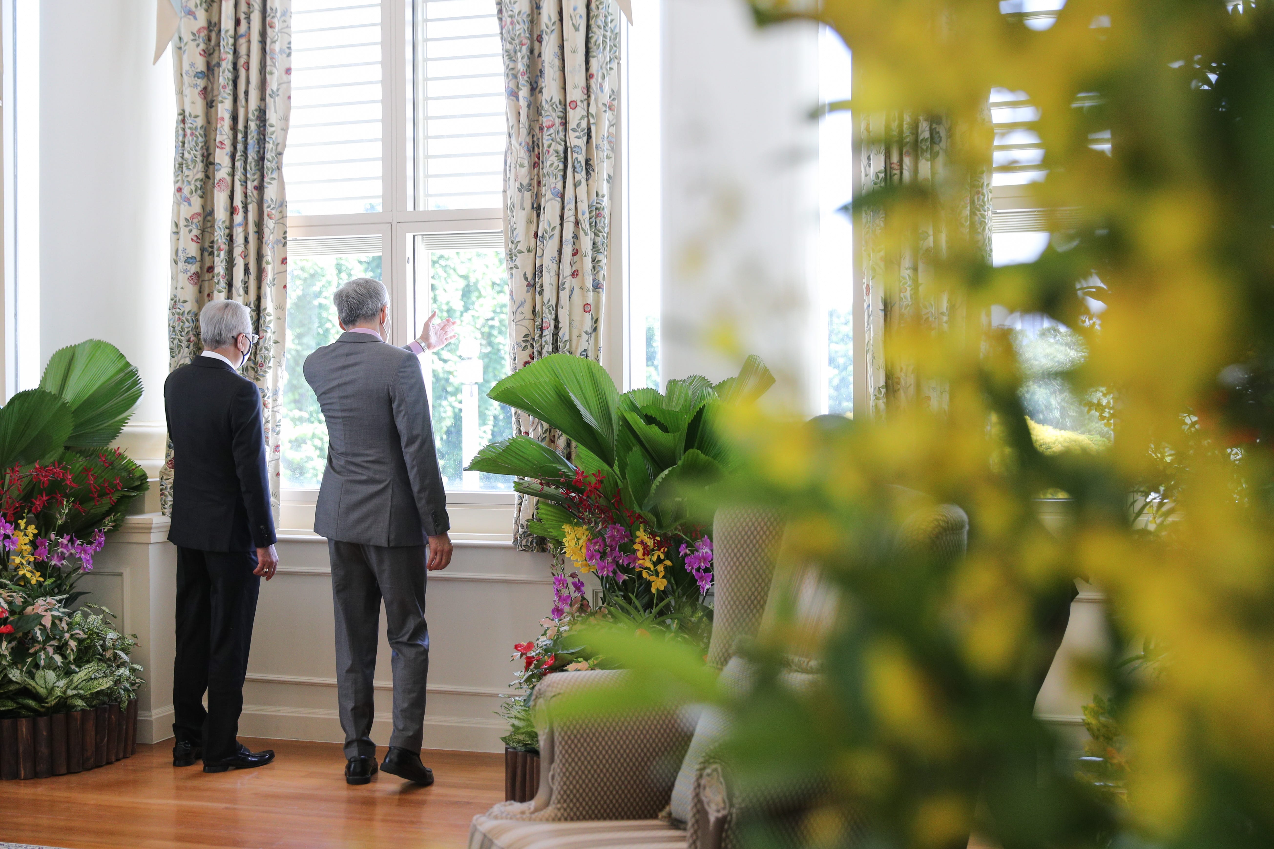 Two men in suits stand by a window with floral curtains, near large potted flowers.