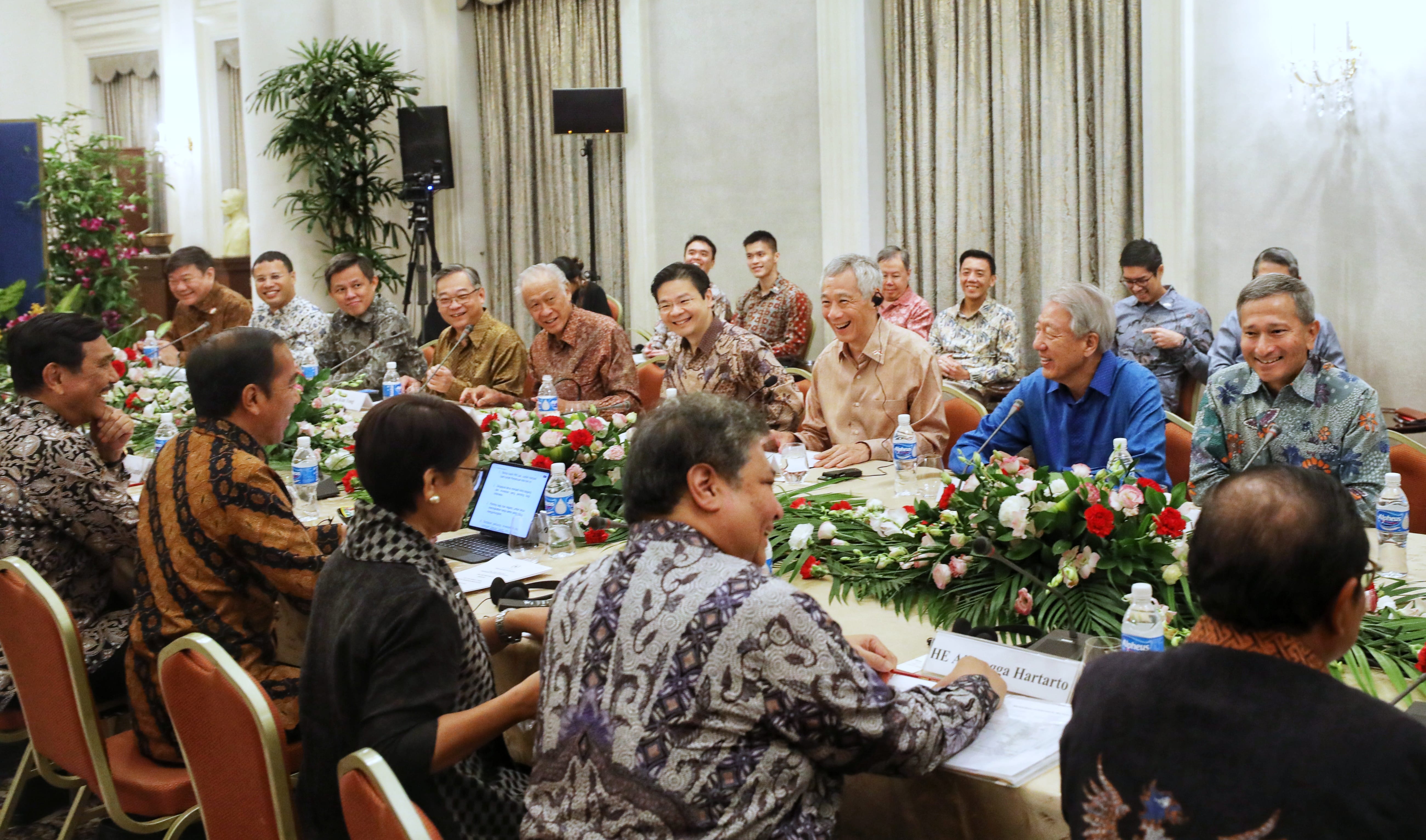 Group seated at long table with flowers and water bottles, some wearing batik-patterned shirts.