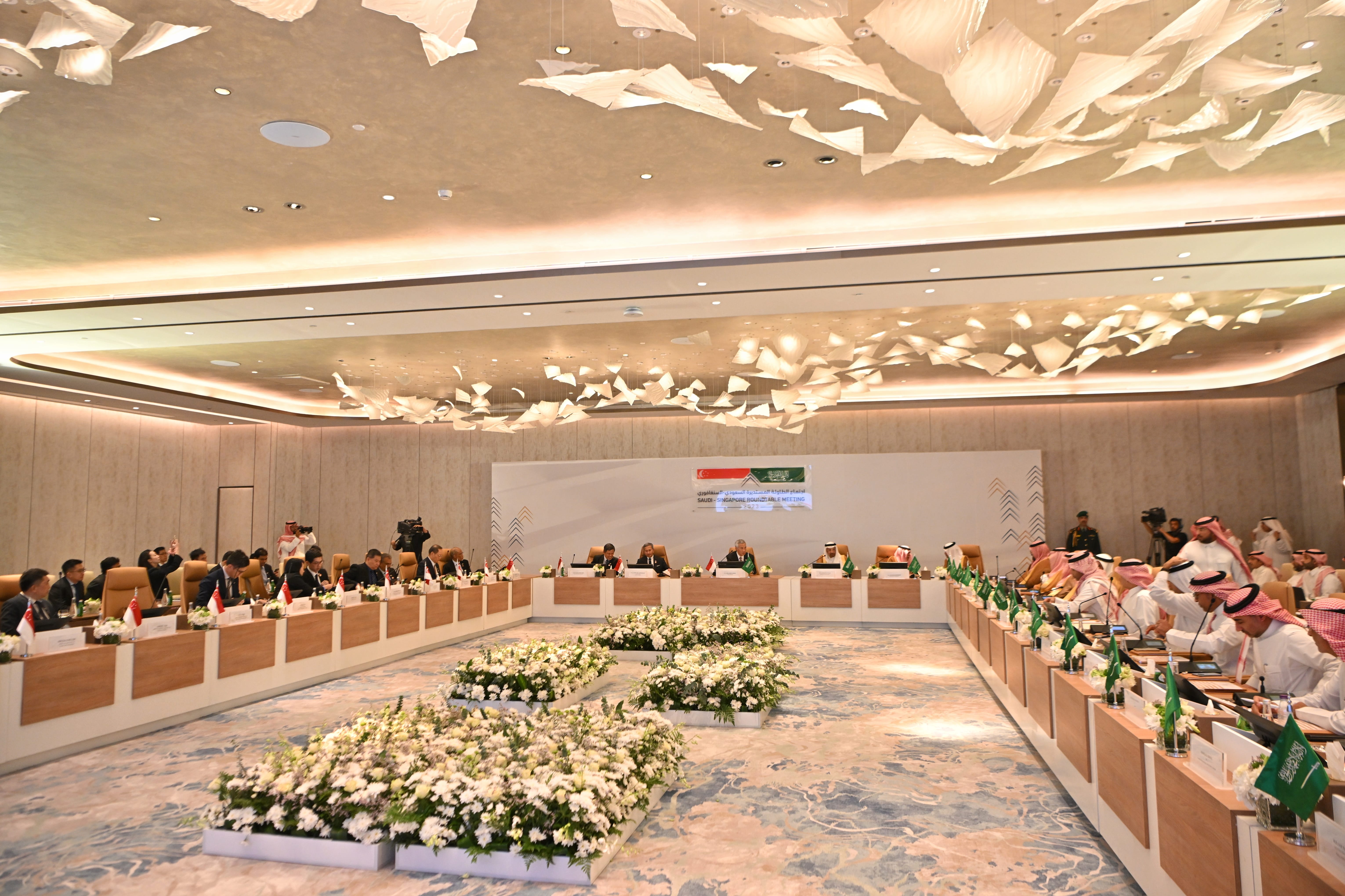 Conference room with people seated at tables, Saudi and Singapore flags displayed.