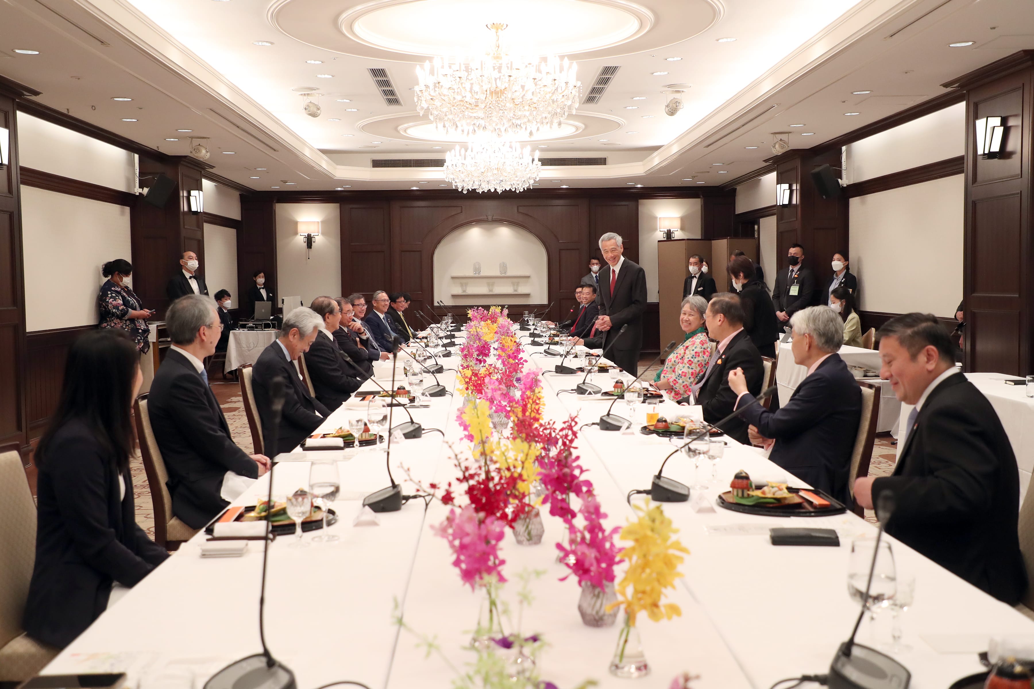 People seated at long table with floral centerpiece, microphones, meal service, crystal chandelier.