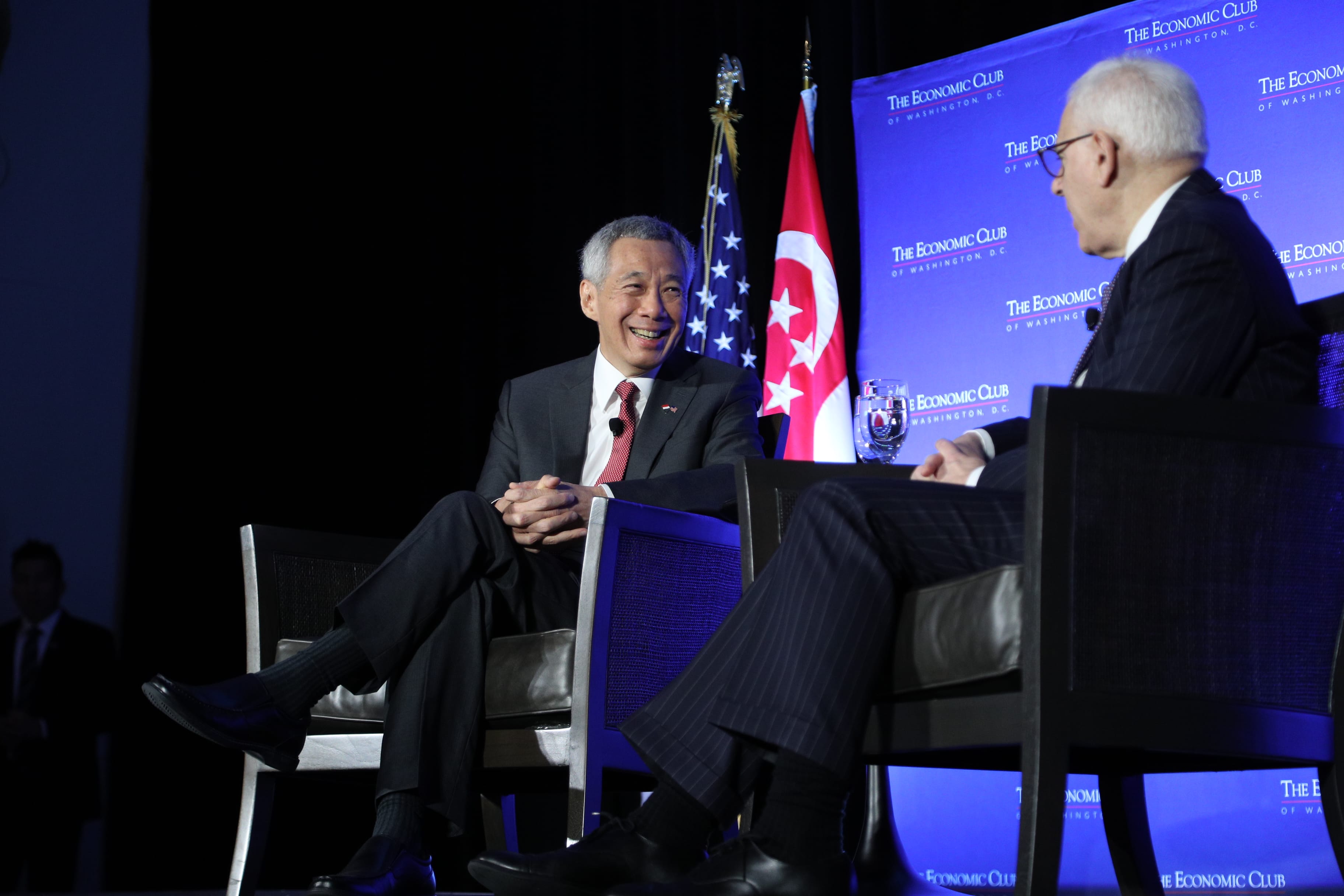 Lee Hsien Loong sits, smiling, in dark suit and red tie with US and Singapore flags behind.