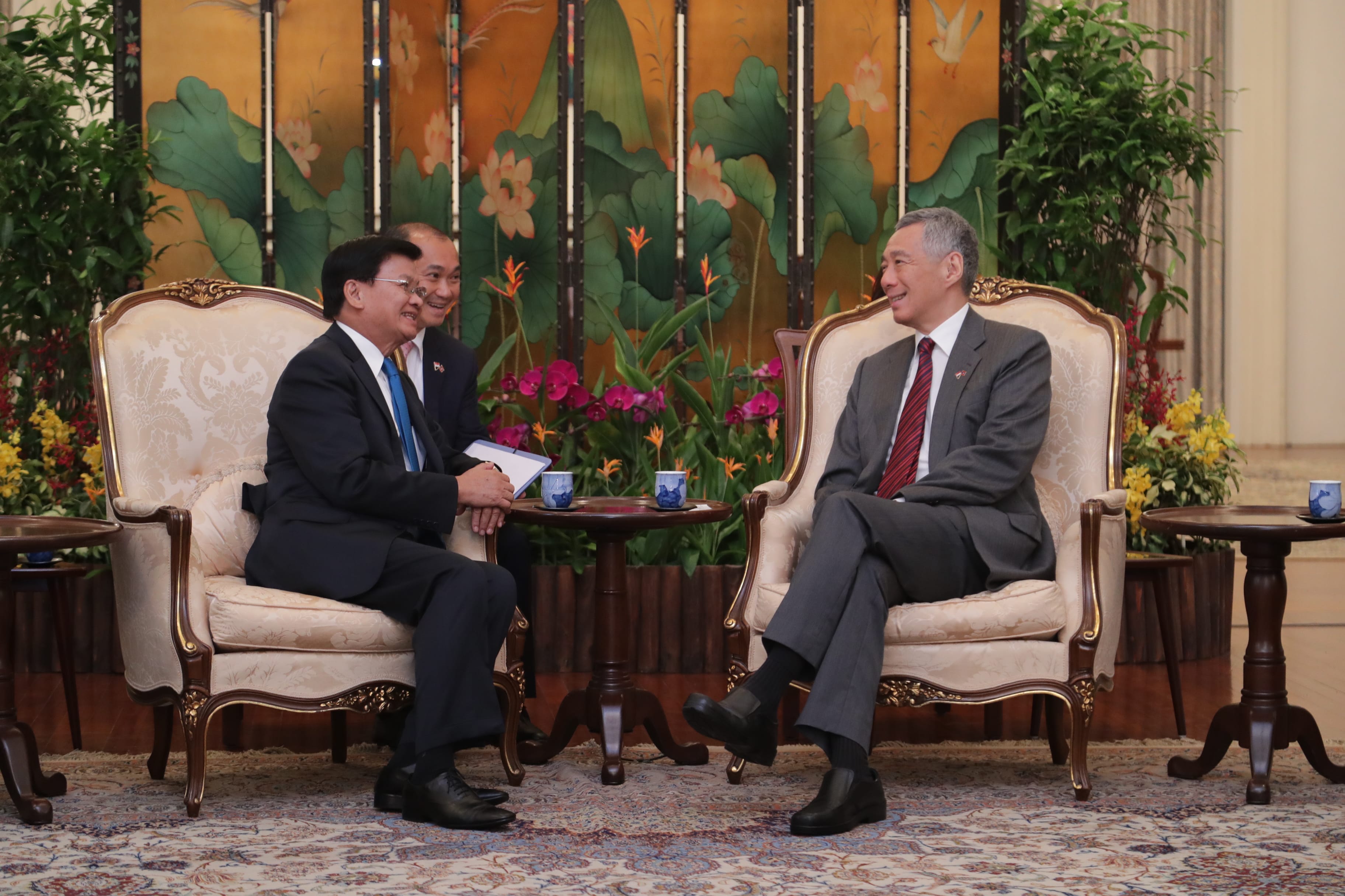 Two men in suits seated in ornate chairs converse, with an Asian floral screen behind them.
