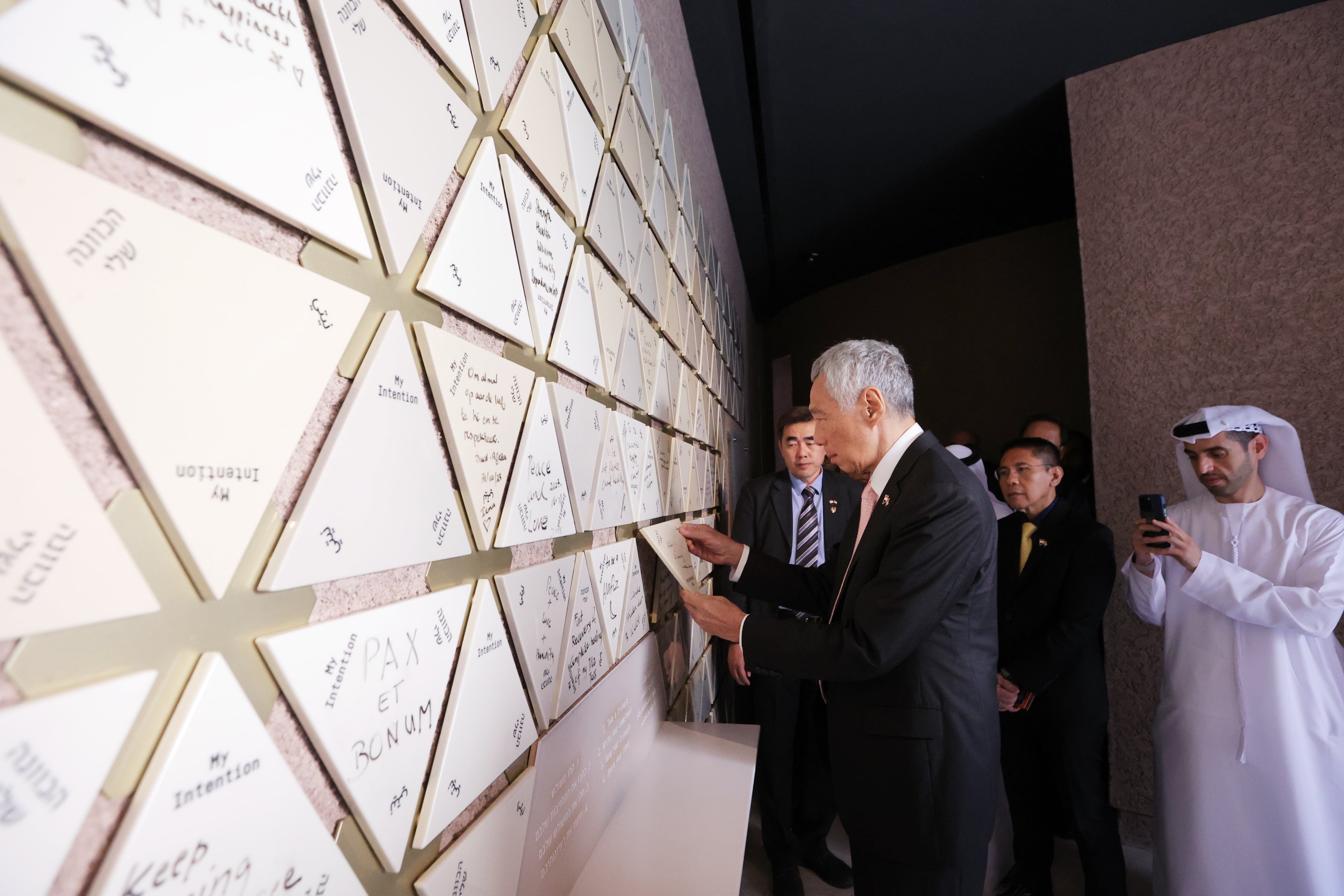 Man in suit examines wall of triangular tiles with written intentions in multiple languages. Other men stand nearby.