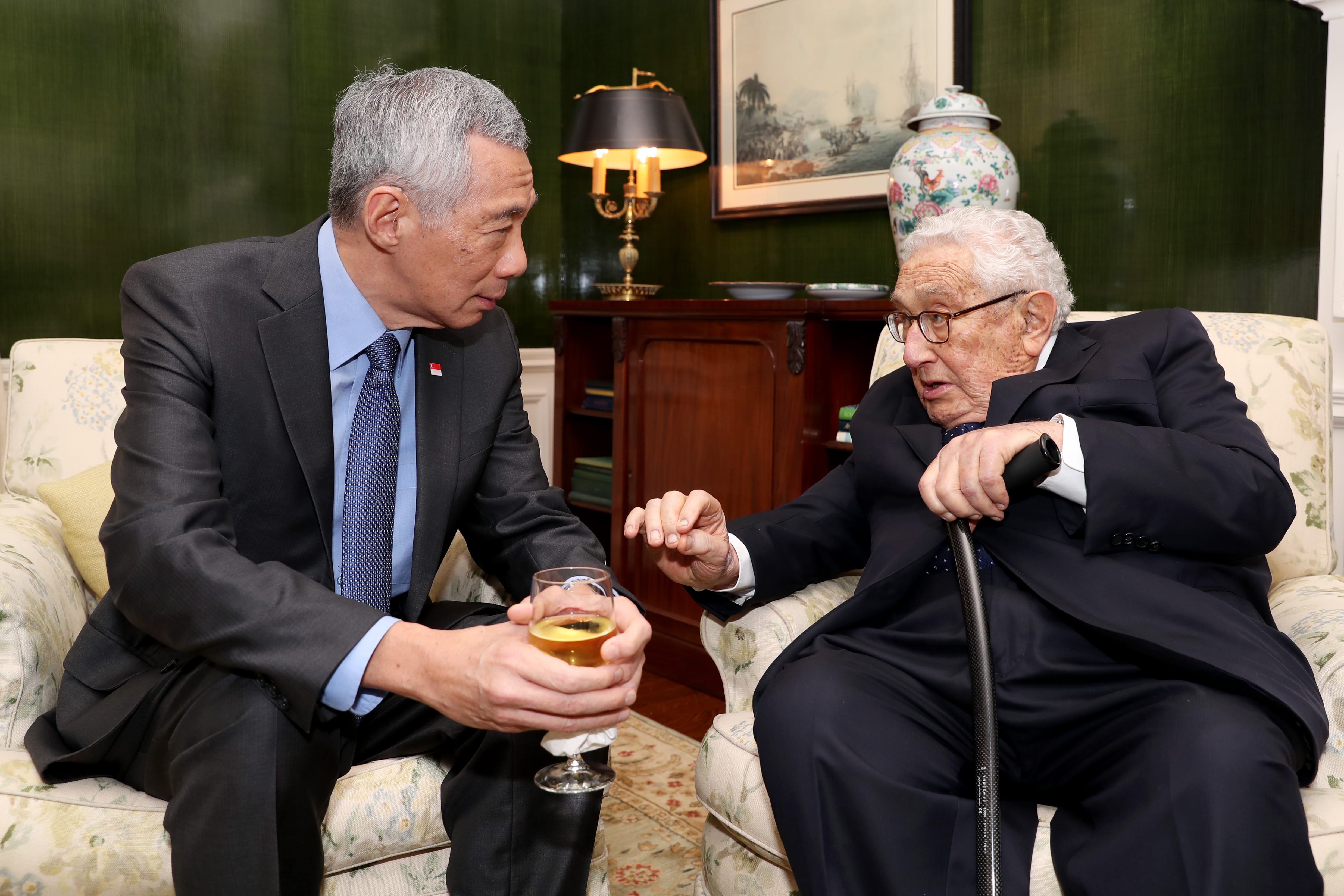 Lee Hsien Loong holding a drink, speaking with Henry Kissinger, who holds a cane.