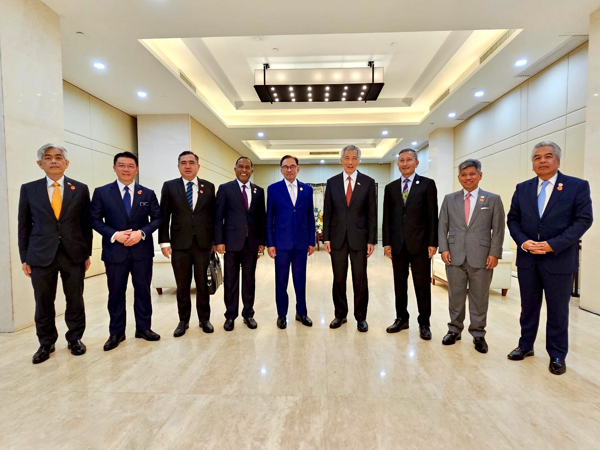 Nine people in business suits standing in a row indoors, with a light marble floor.