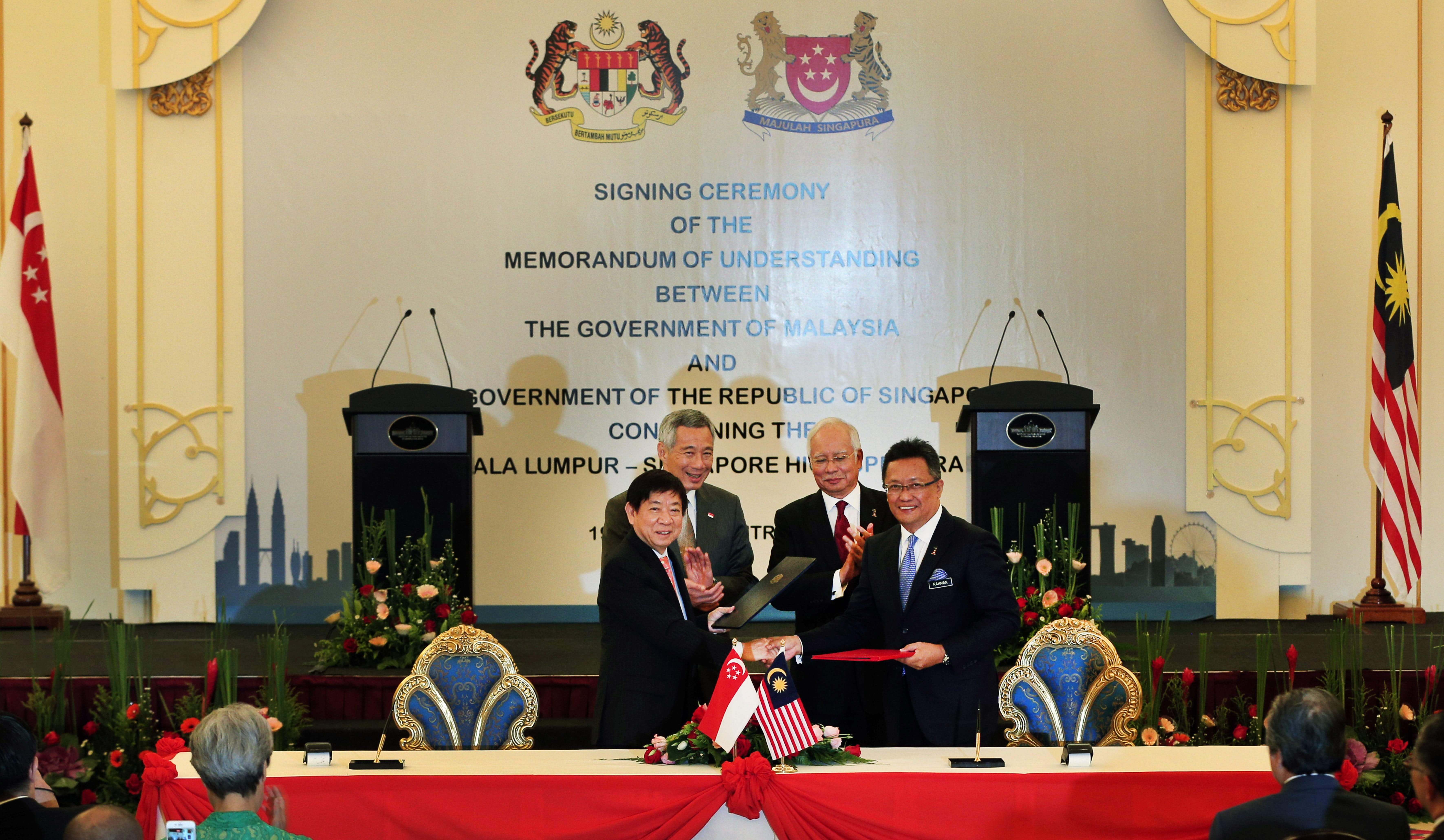Malaysia/Singapore signing ceremony: men shaking hands over documents, flags, and table with floral decor.
