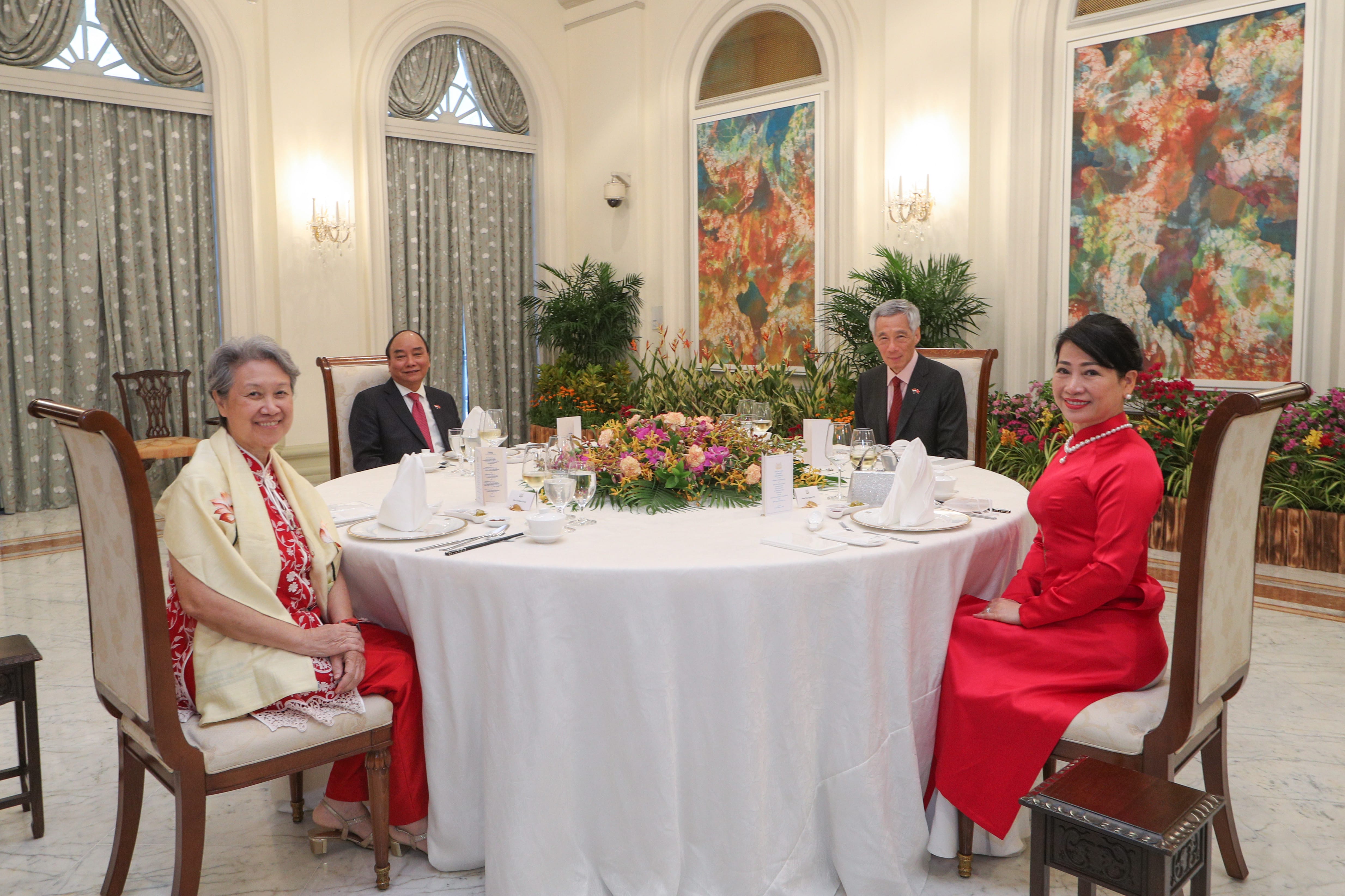 Four people seated at a round table set with dinnerware, surrounded by plants, flowers, and abstract paintings.