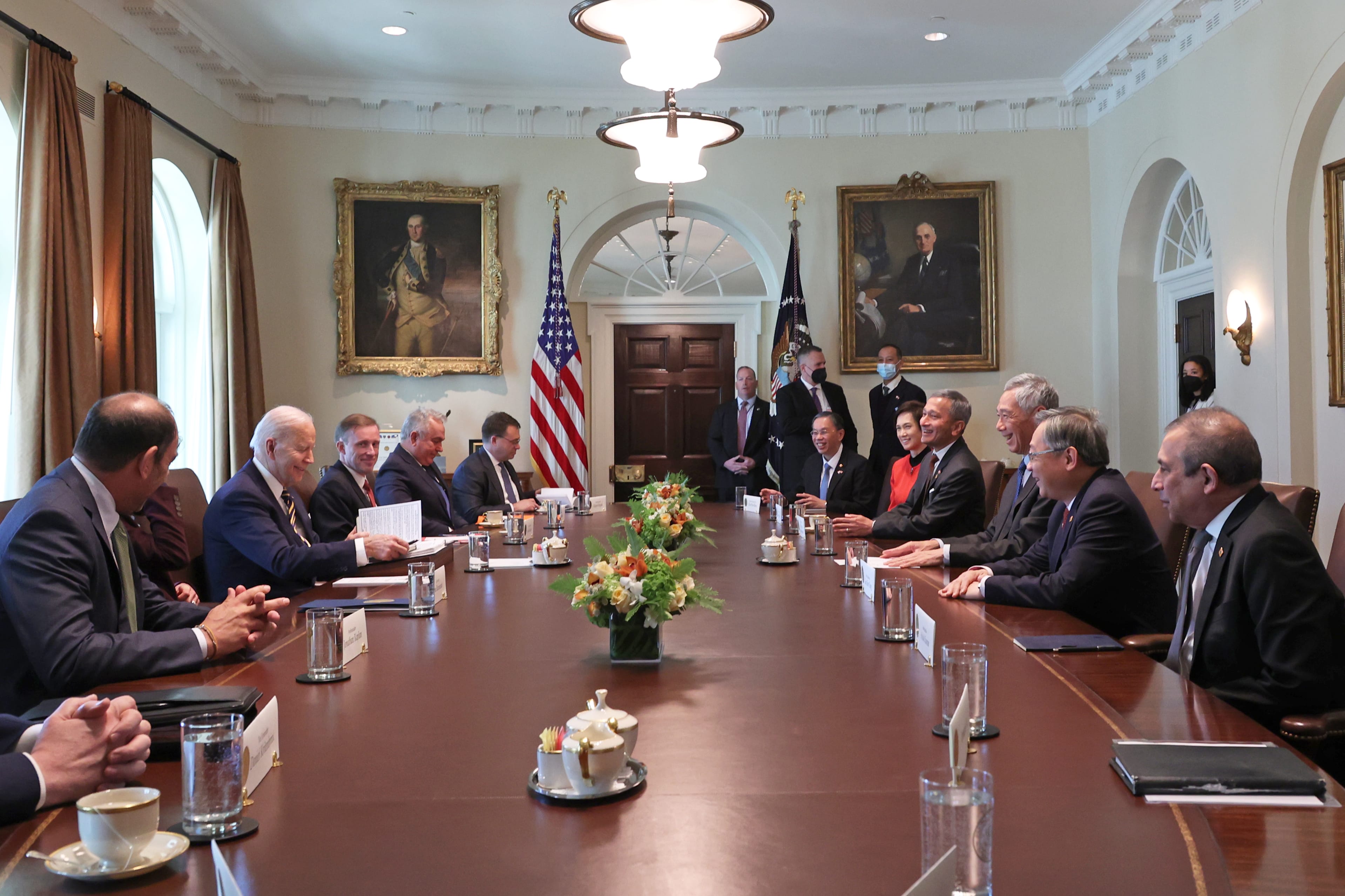 Oval Office meeting: Joe Biden and others seated at a long table with flags and floral arrangements.