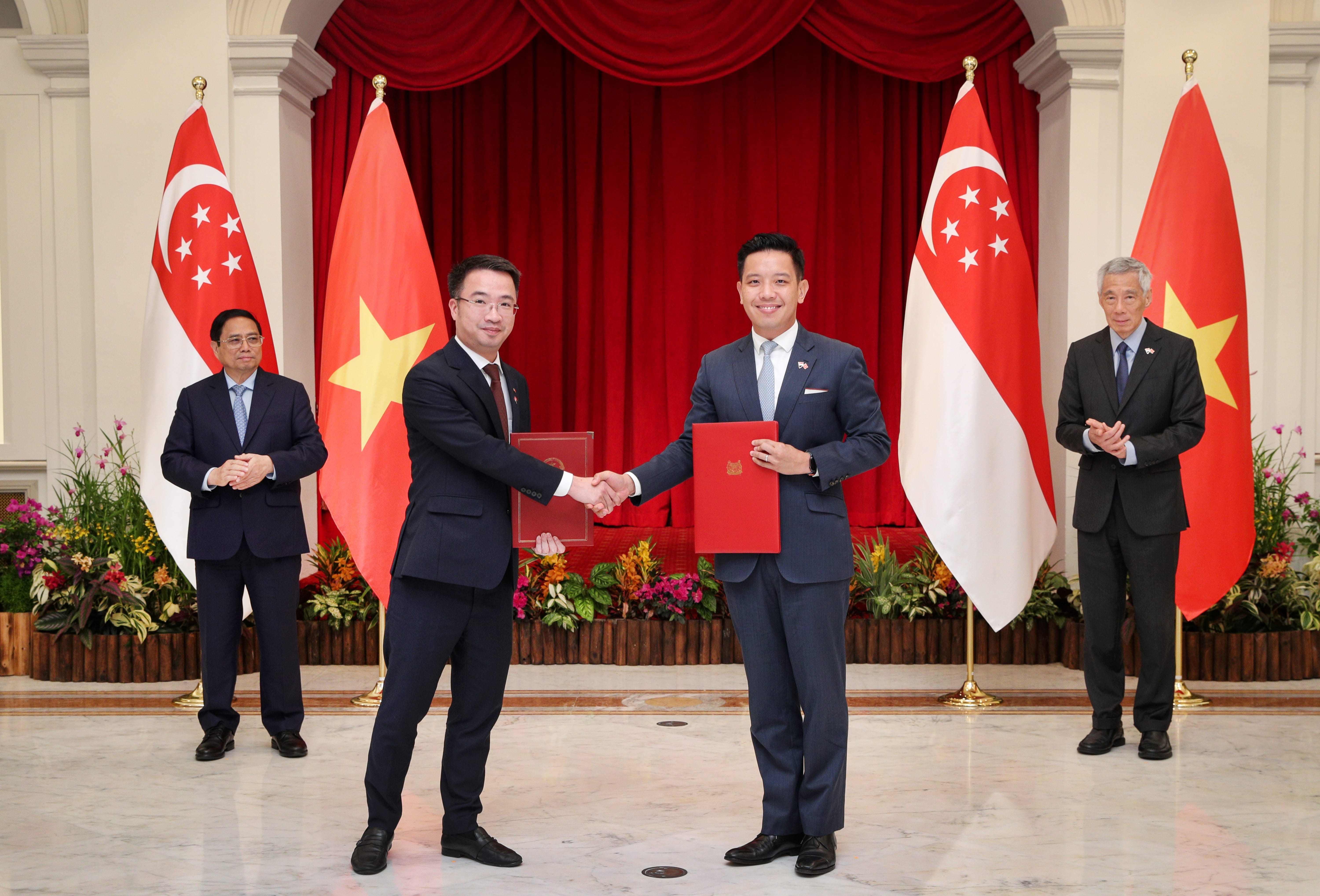 Two men in suits shaking hands, each holding a red folder, flags of Singapore and Vietnam in background.