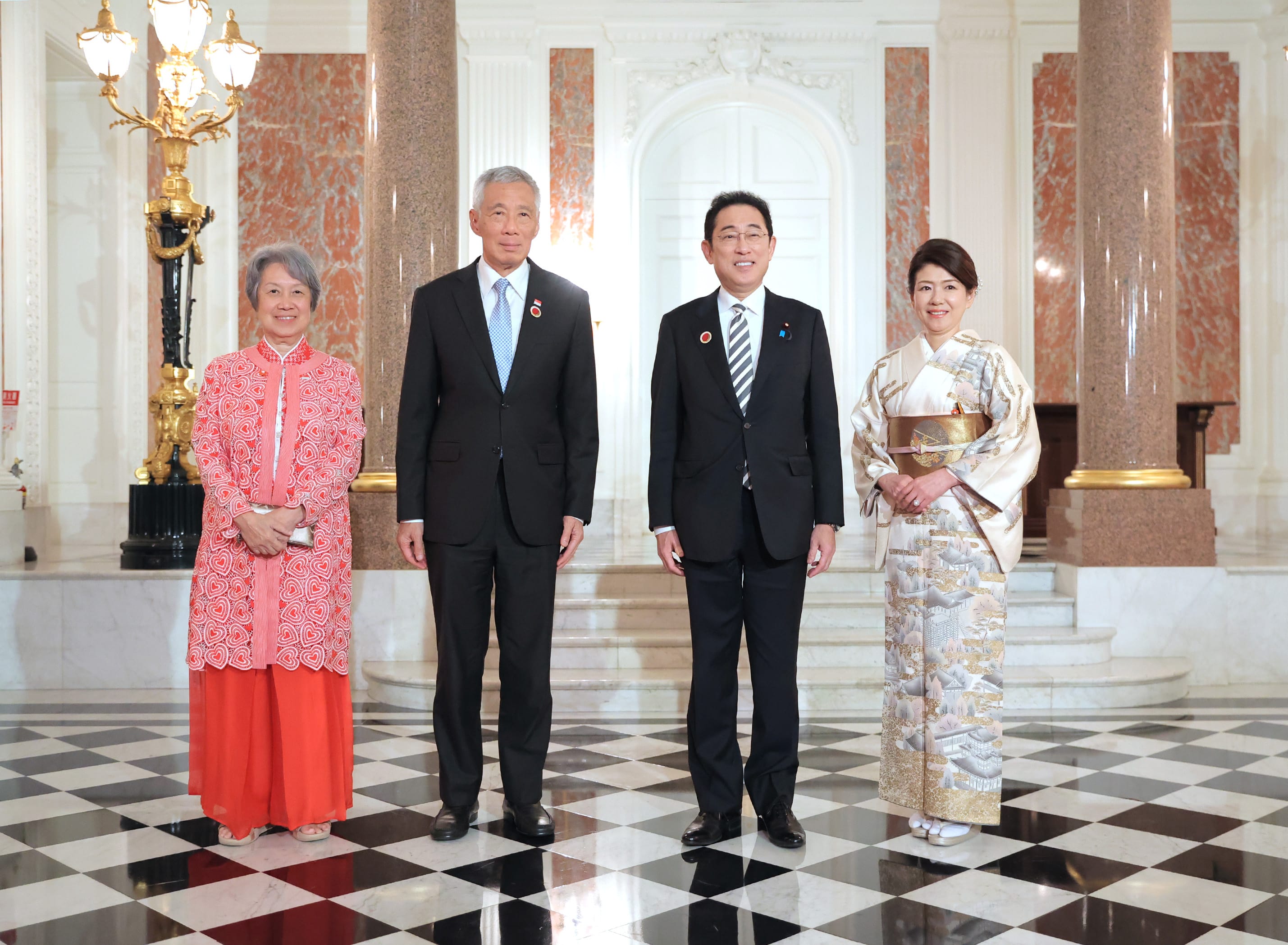 Four people stand on a black and white tile floor, facing forward, with marble pillars behind them.