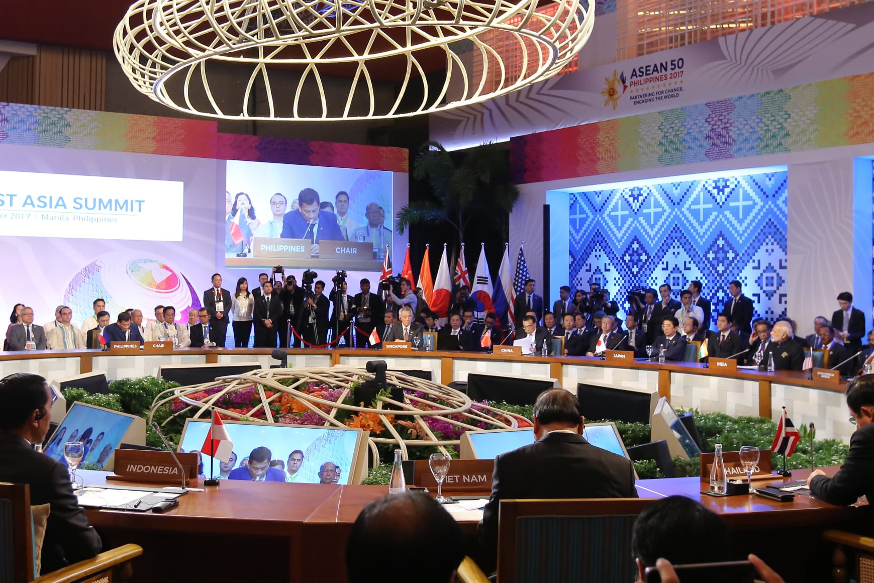 Heads of state at East Asia Summit seated at tables with country names and flags. Monitors show speaker.
