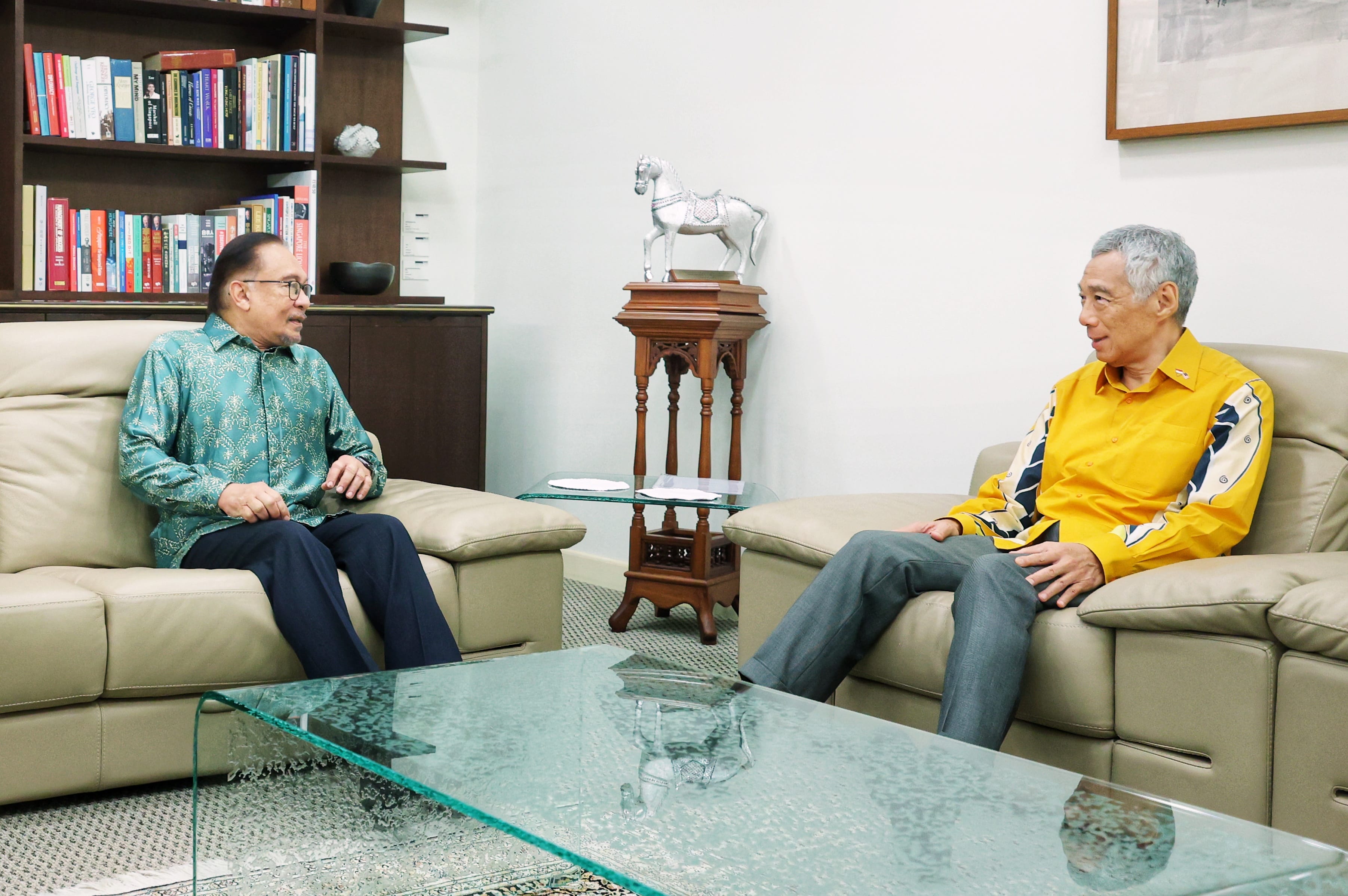 Anwar Ibrahim and Lee Hsien Loong sit on a sofa in a room with a glass table, bookcase, and horse statue.