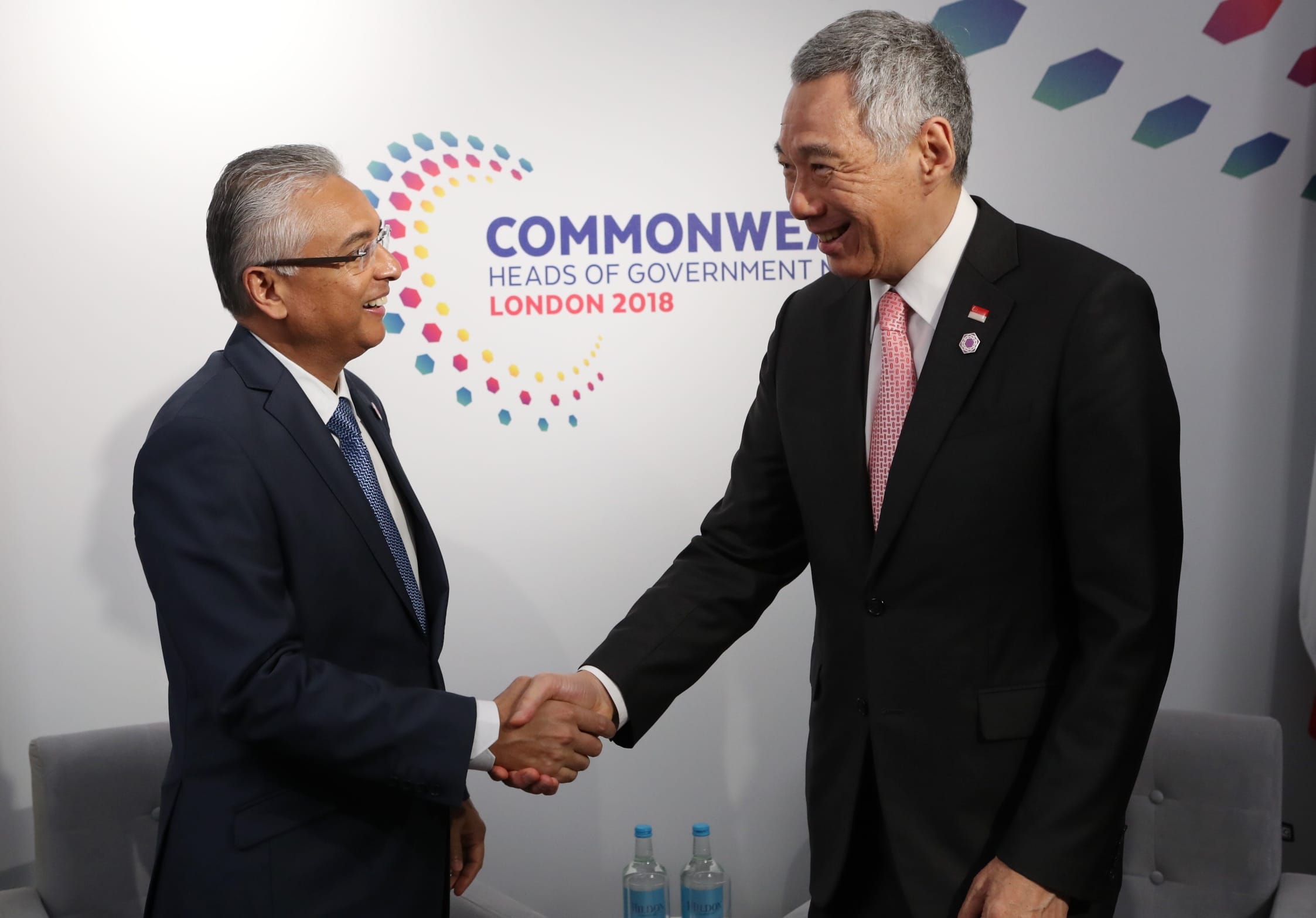 Two men in suits shake hands, Commonwealth Heads of Government logo behind them.