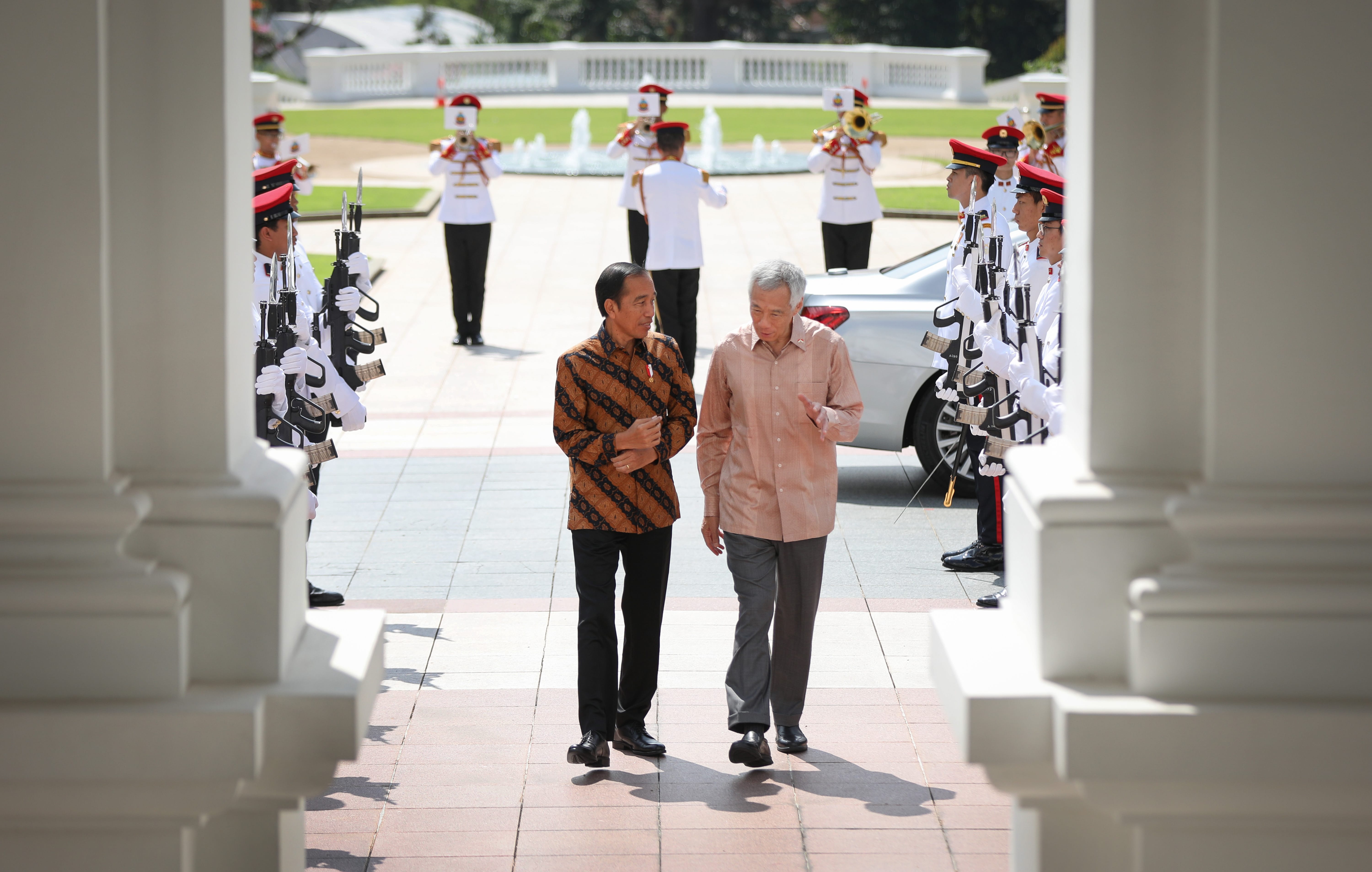 Joko Widodo and Lee Hsien Loong walk amid guards and musicians in white uniforms with red trim.