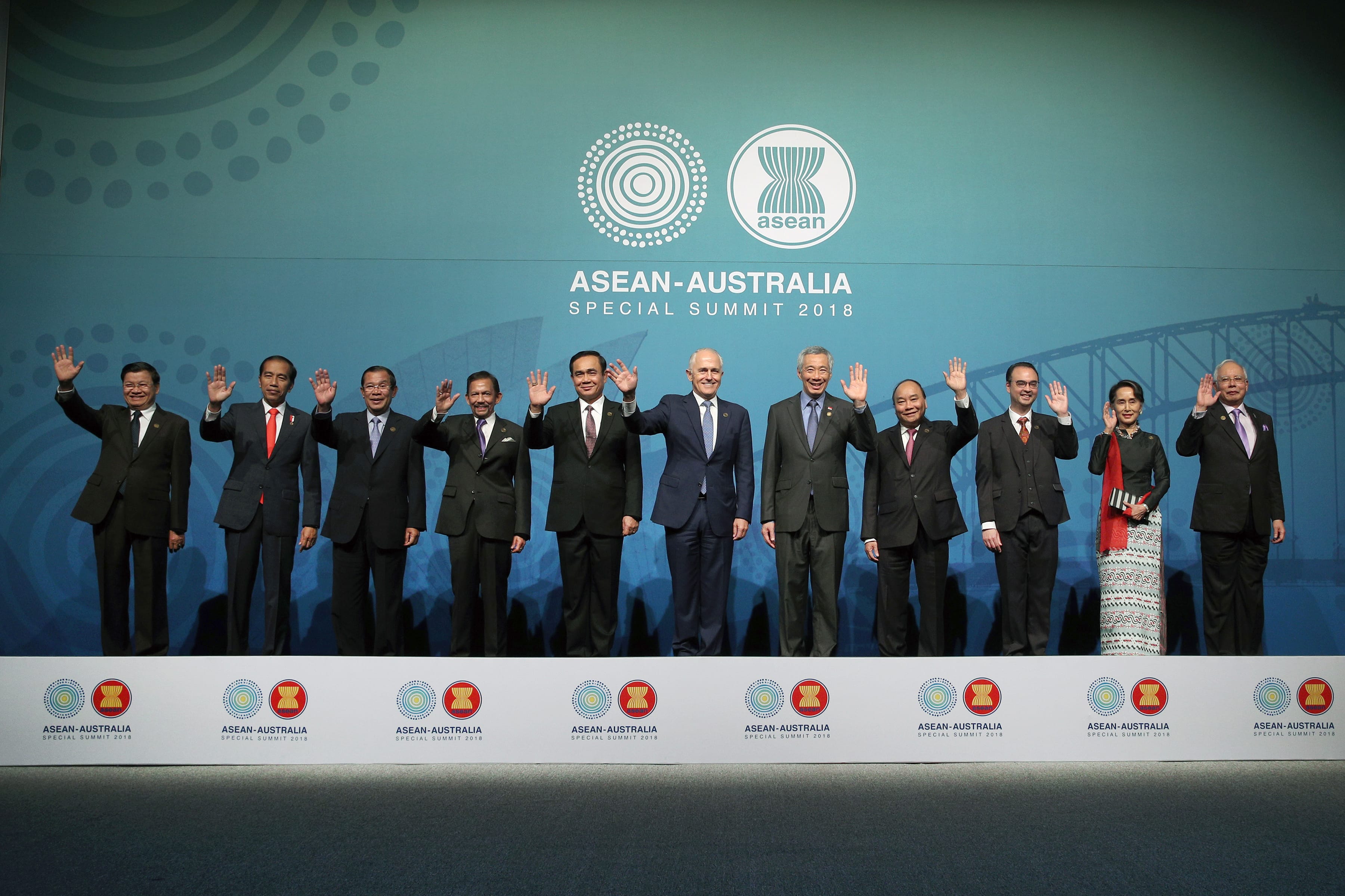 Eleven leaders on a stage with "ASEAN-Australia Special Summit 2018" displayed on a screen behind them, waving.