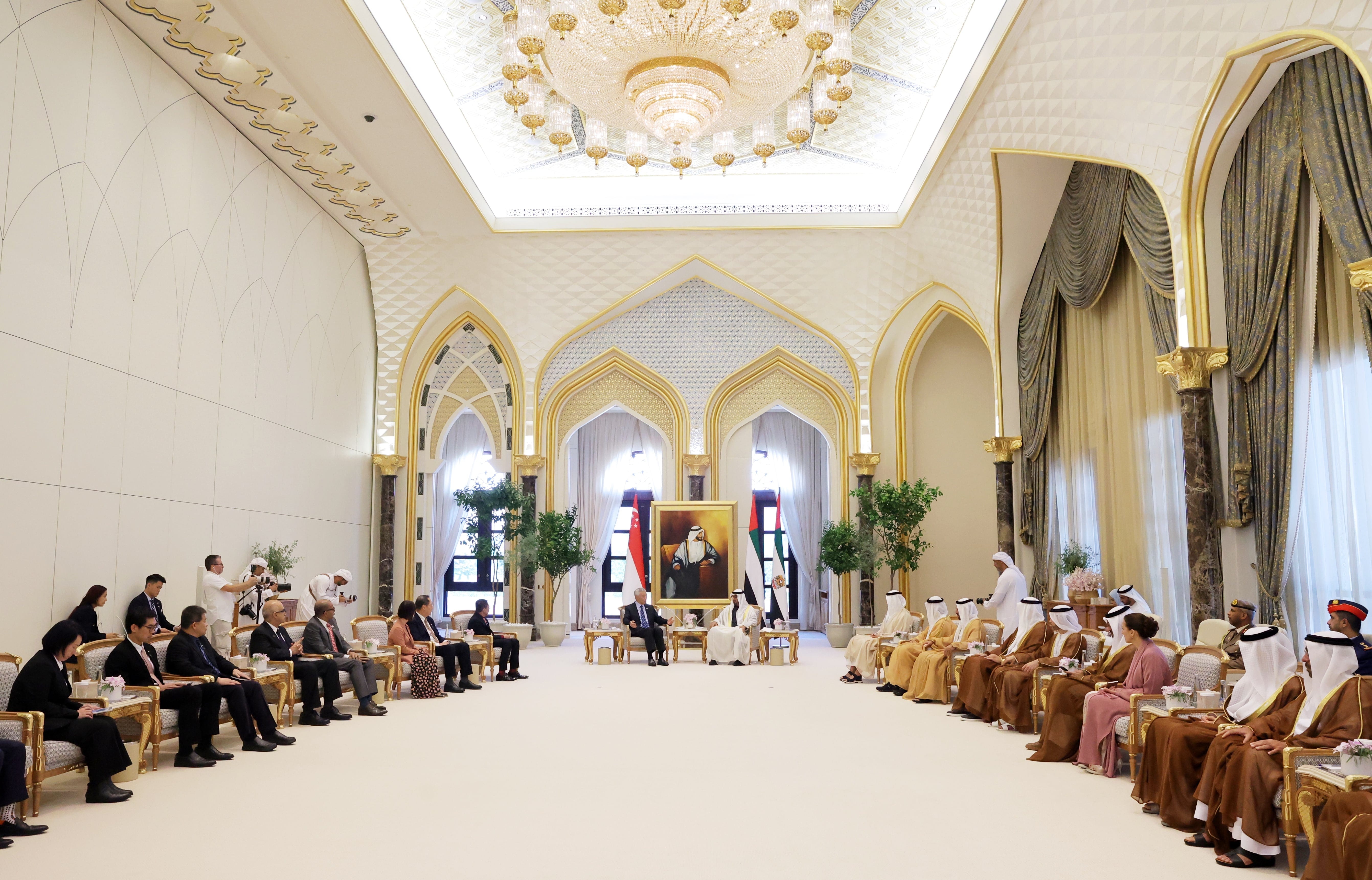 Large ornate room with groups of people seated facing each other. Chandeliers overhead.
