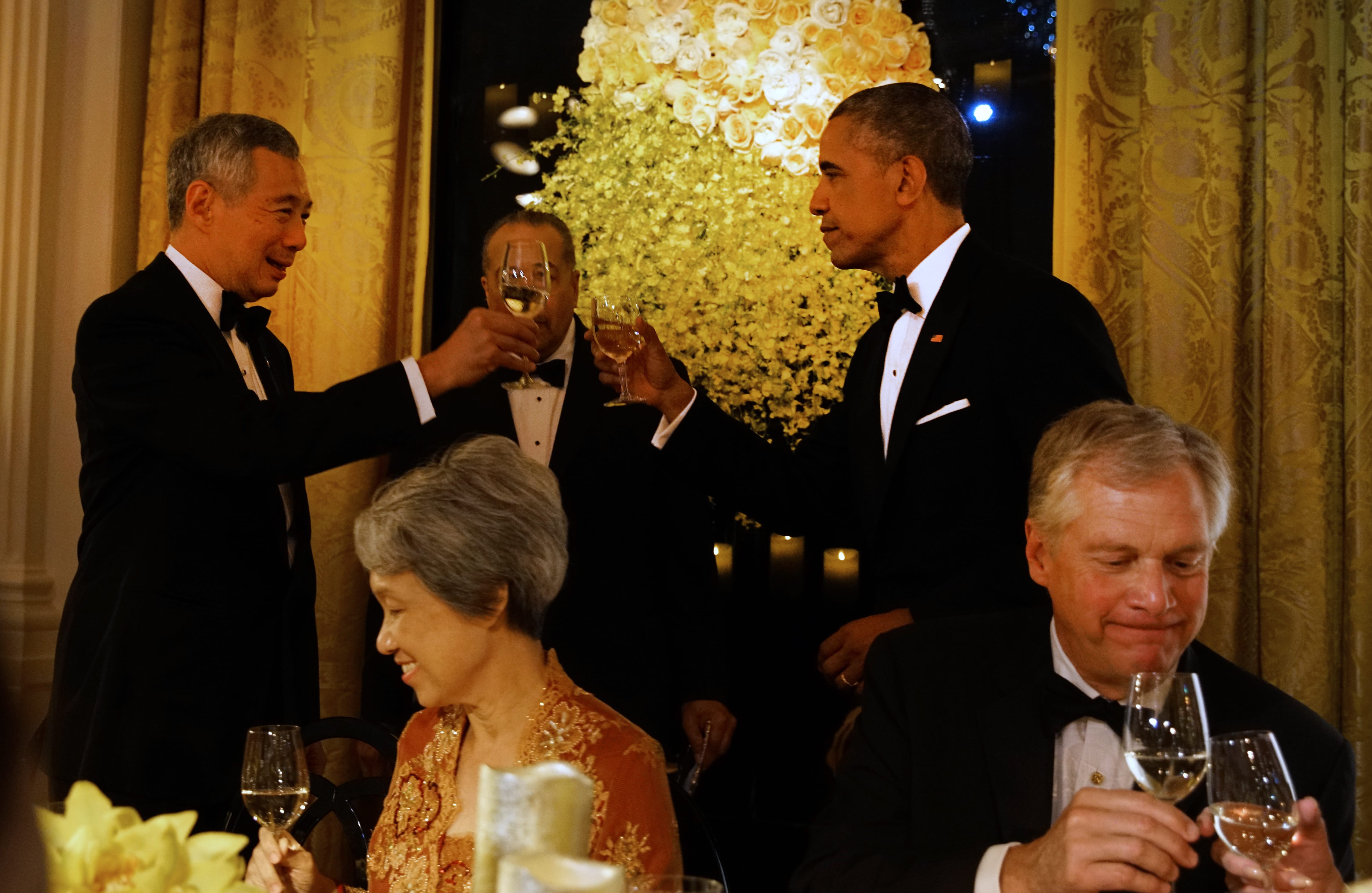 Barack Obama & Lee Hsien Loong in tuxedos toast with champagne. Floral centerpiece backdrop.