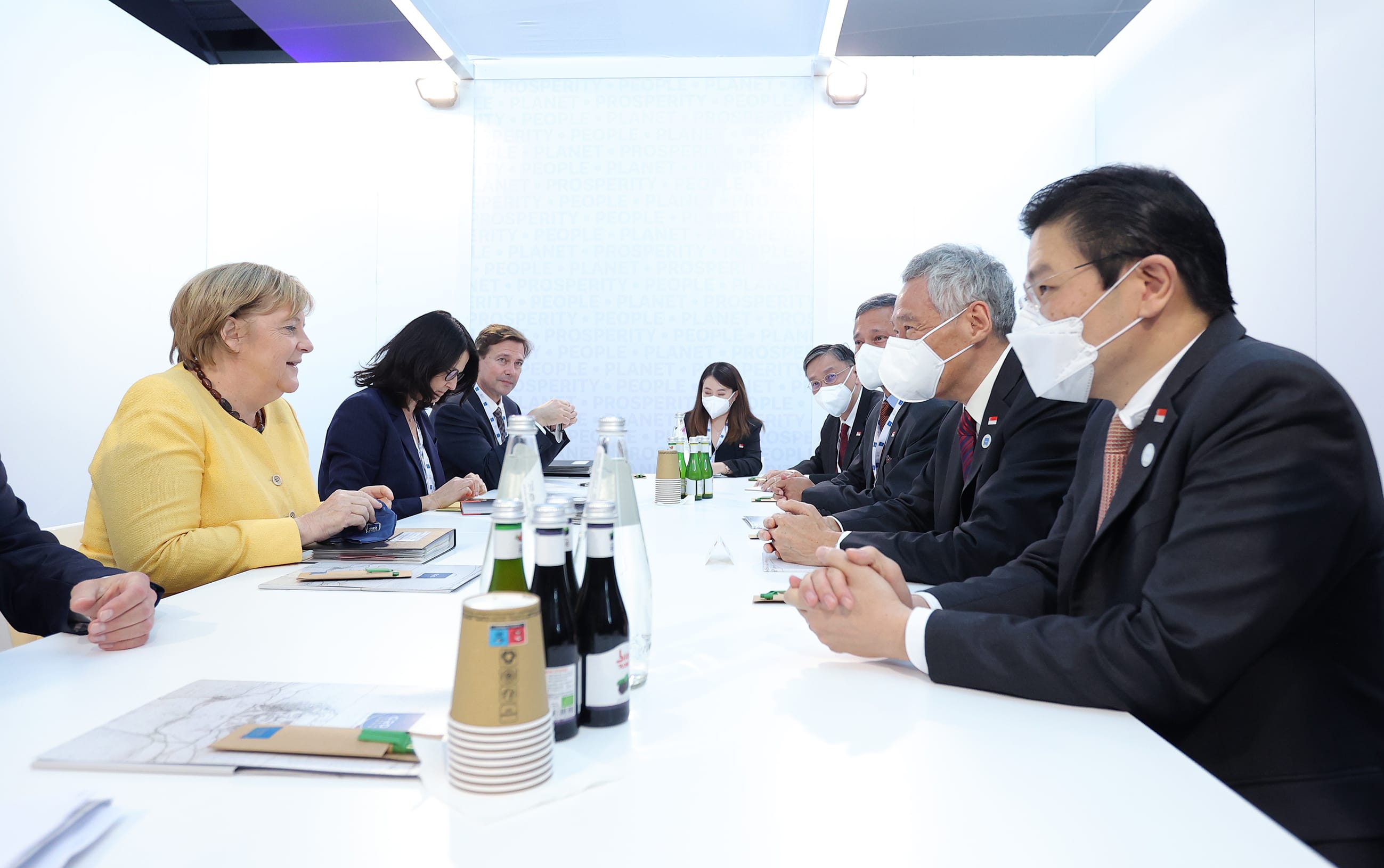 People at a table, some wearing face masks. A woman in yellow, and others in dark suits.