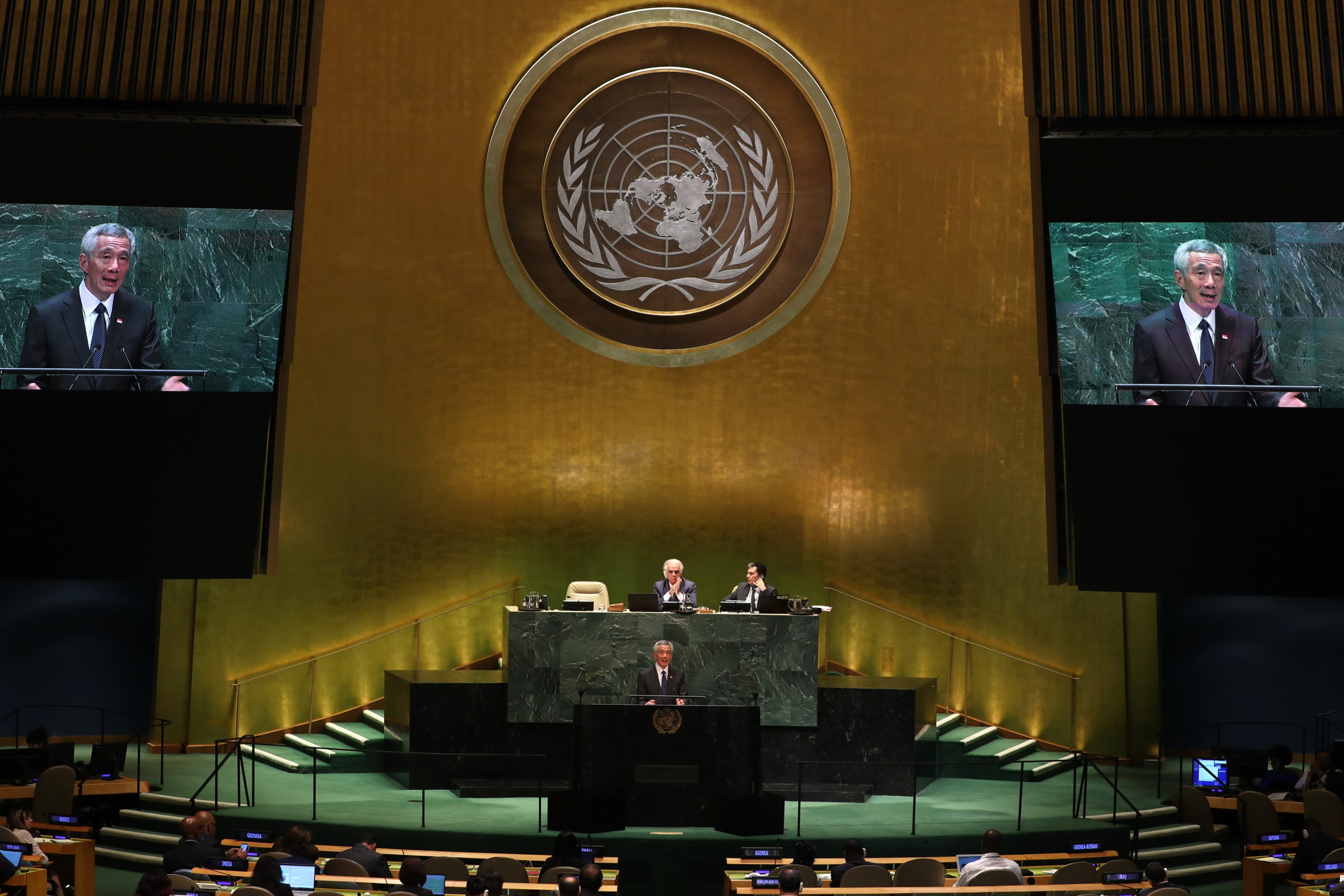 UN General Assembly hall with speakers, large screens, audience, and UN logo.