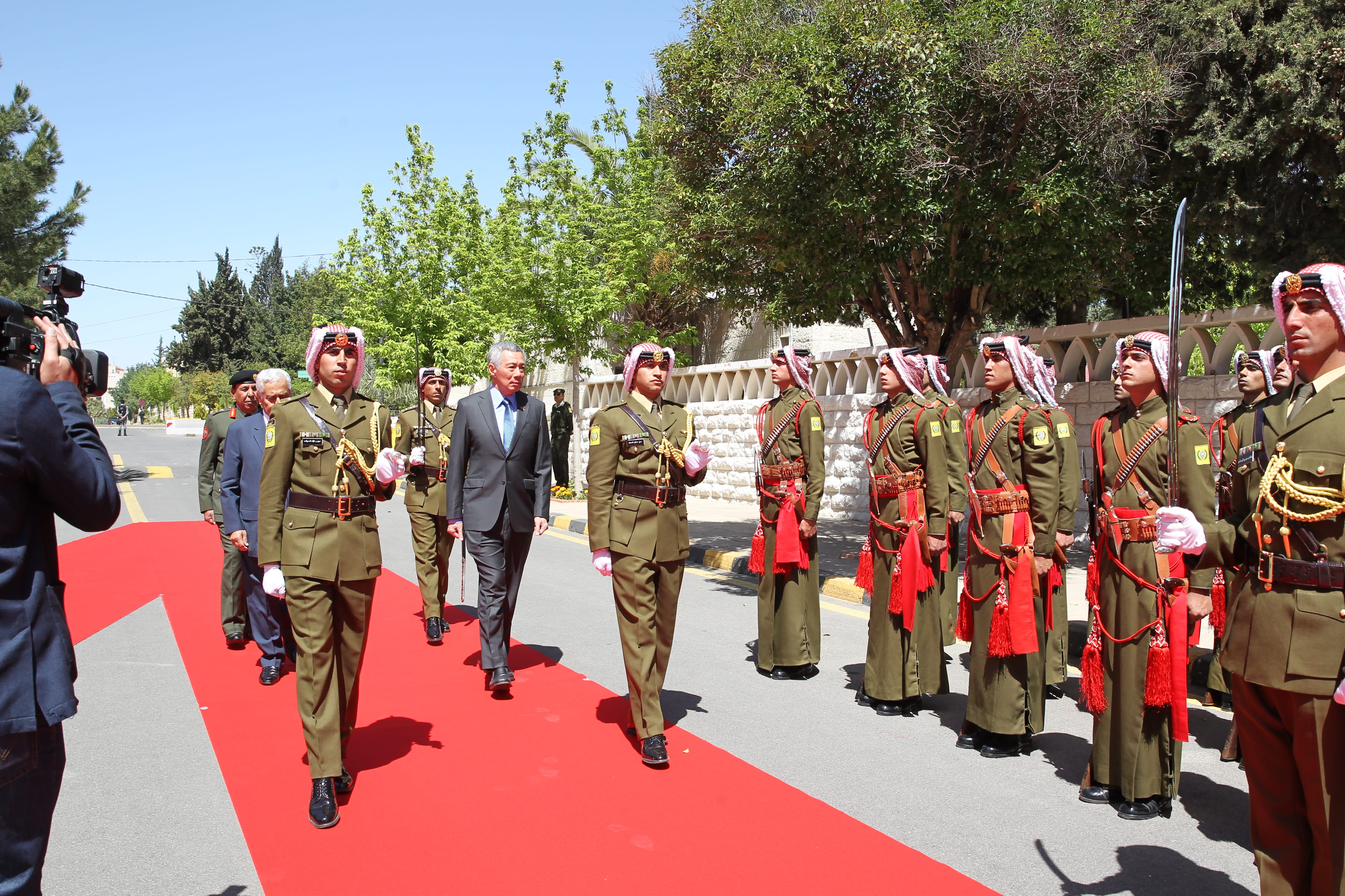 Dignitaries walk red carpet with honor guard in olive uniforms and red-and-white headdresses.