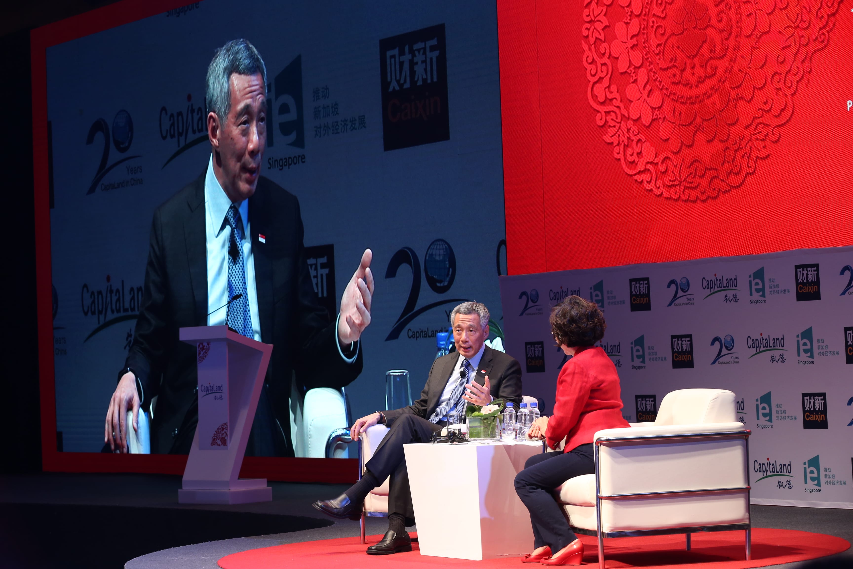 Lee Hsien Loong on screen and on stage with a woman on a stage with branding.
