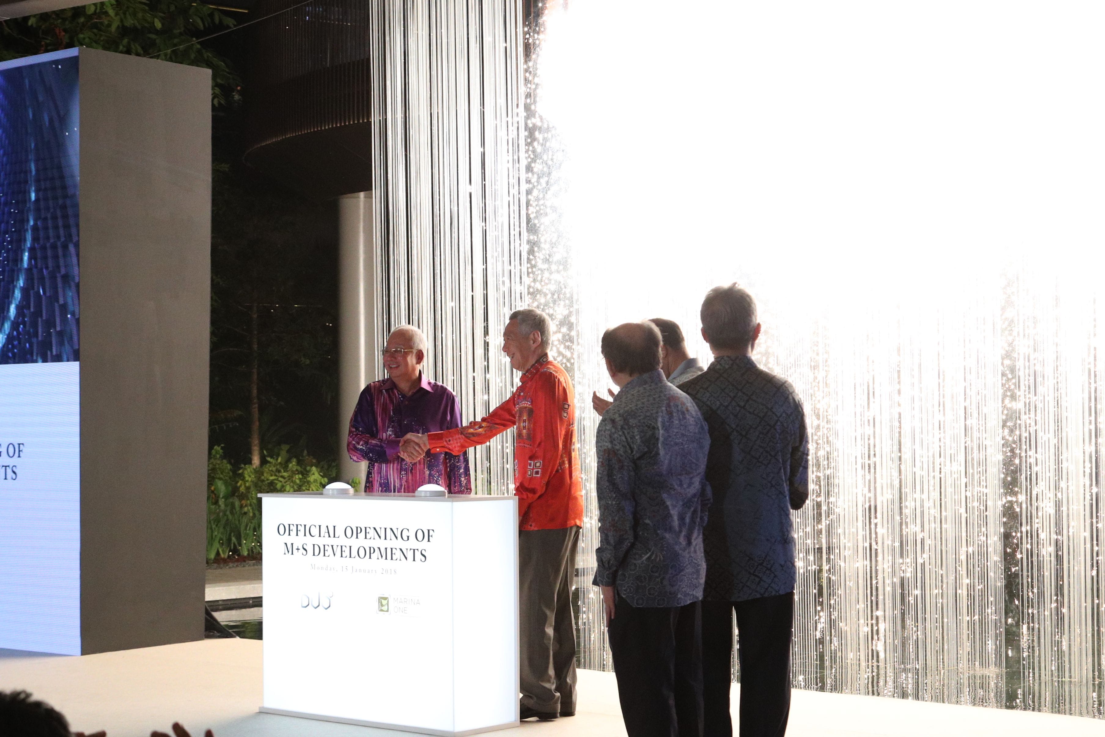 Two men shake hands on a stage with a white light-up banner and decorative light curtain.