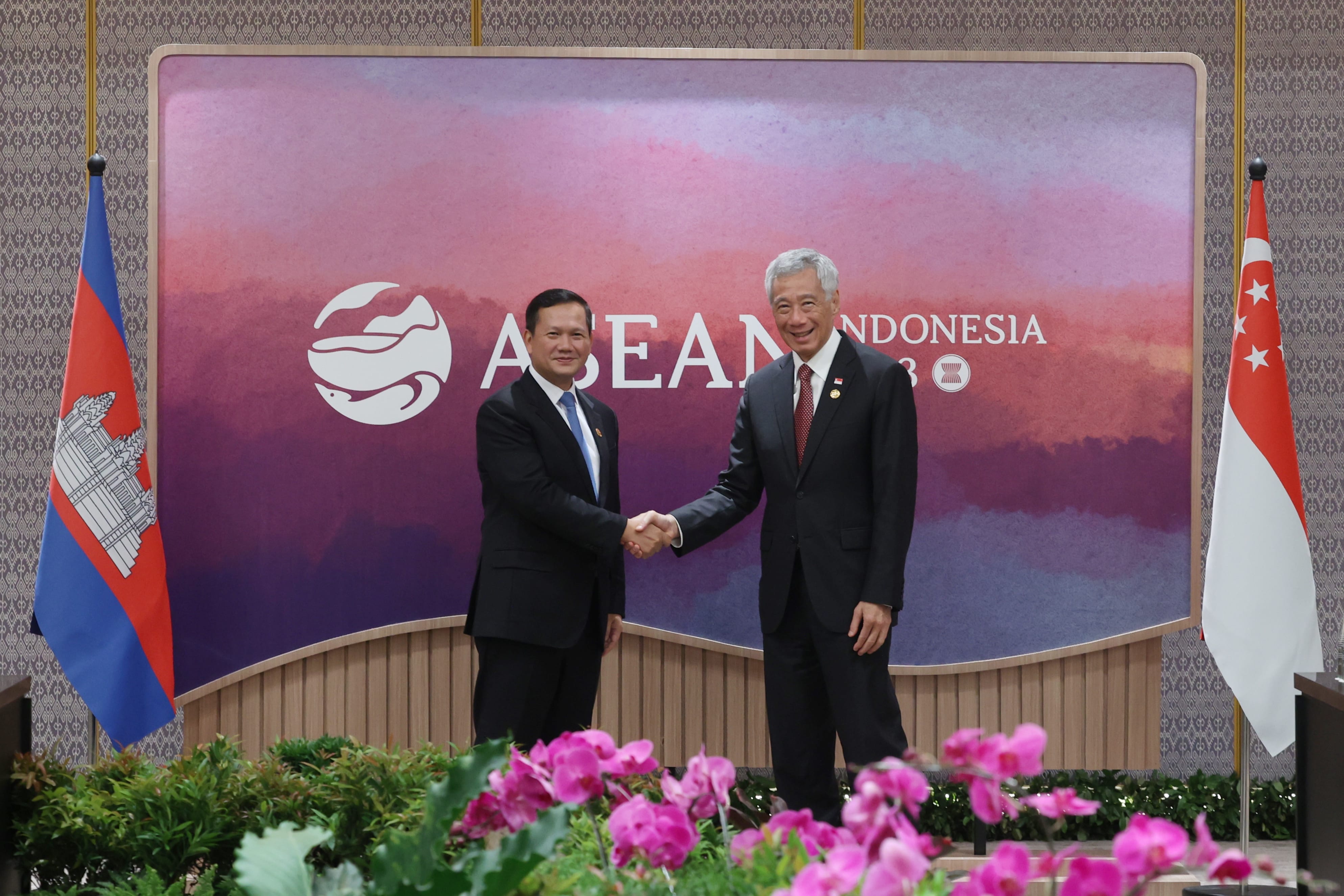 Two men in suits shake hands before an ASEAN backdrop with Cambodia and Singapore flags.