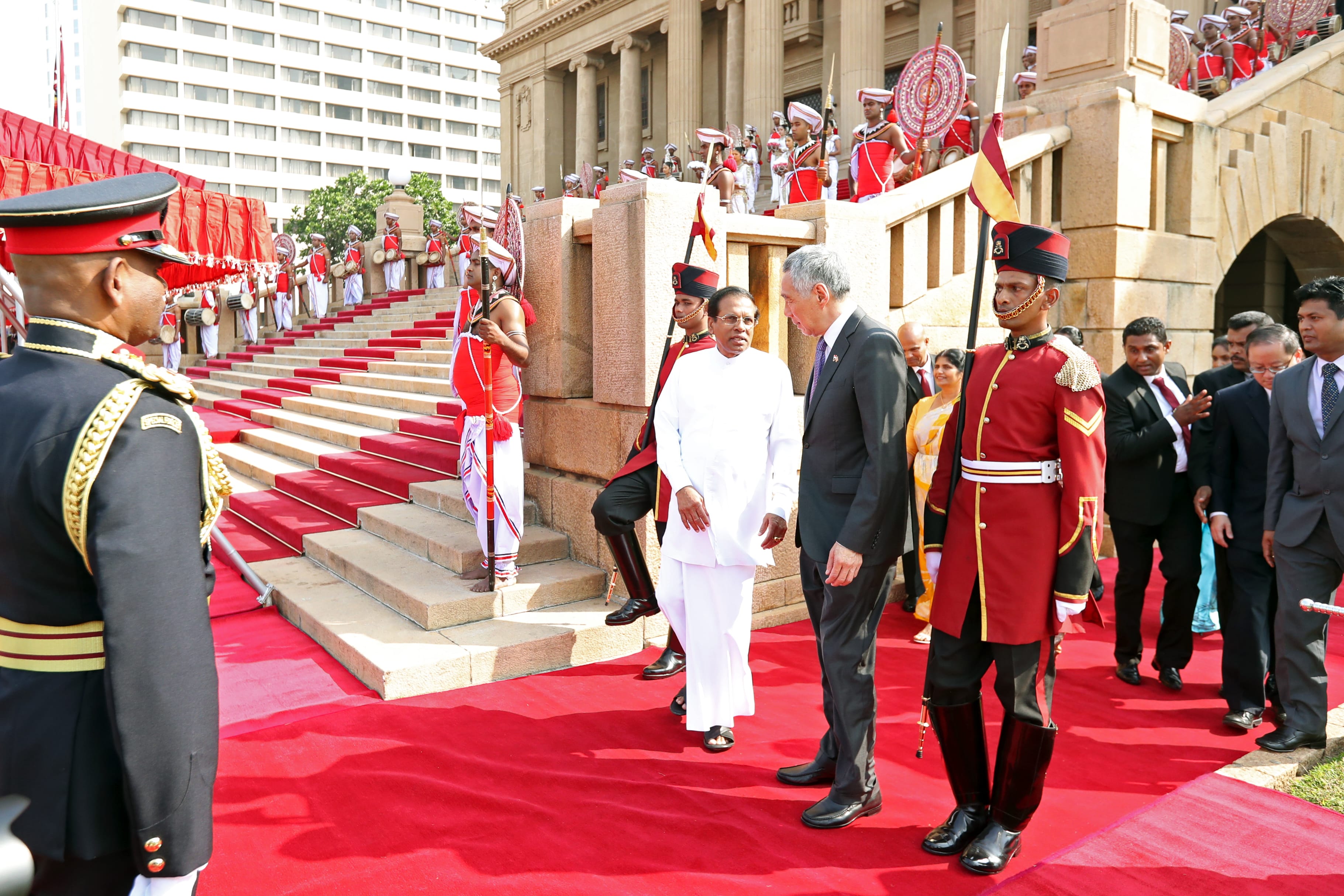 Two men in formal wear on red carpet flanked by guards and musicians on steps of building.