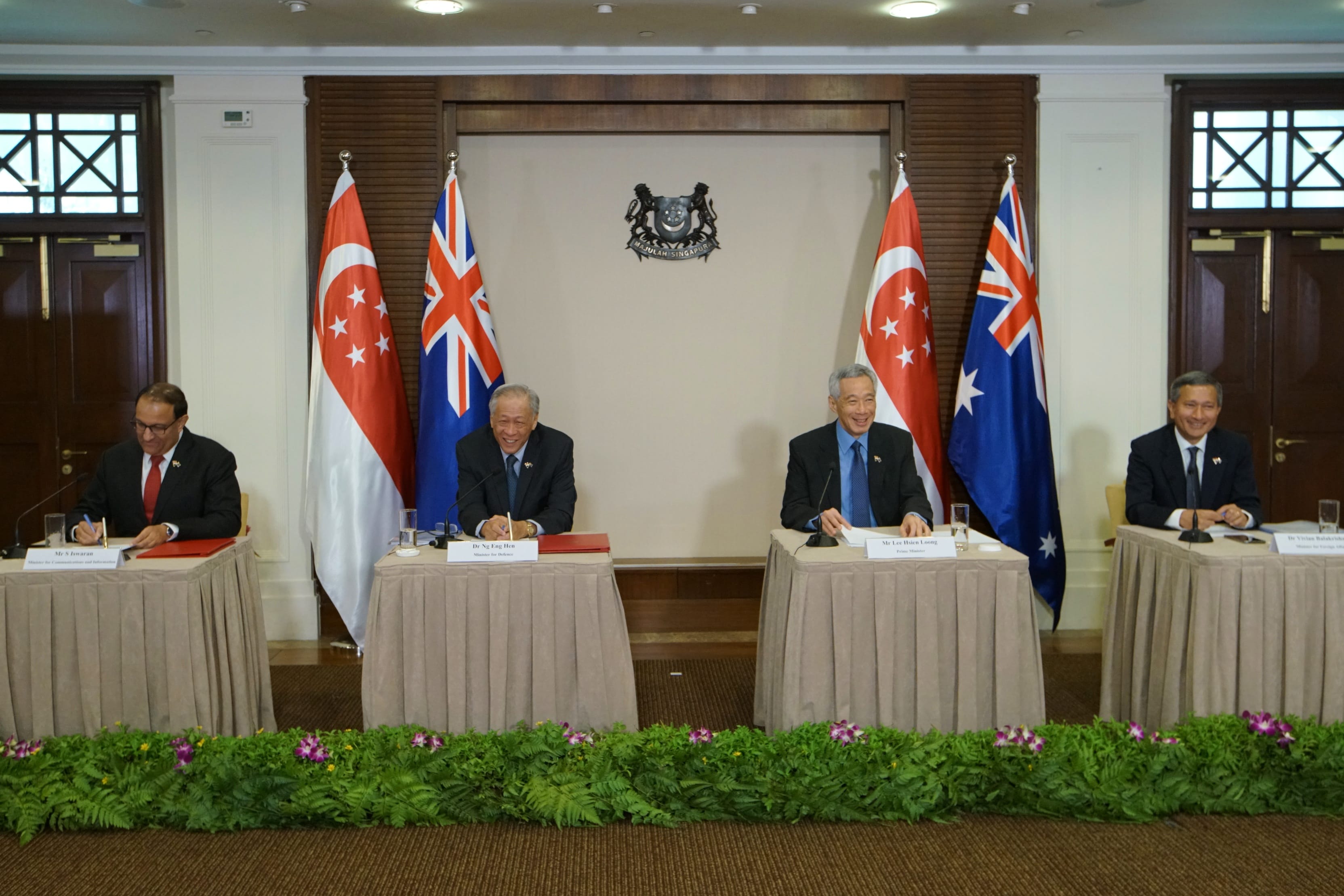 Four people at desks between Singapore and Australia flags, with floral greenery.