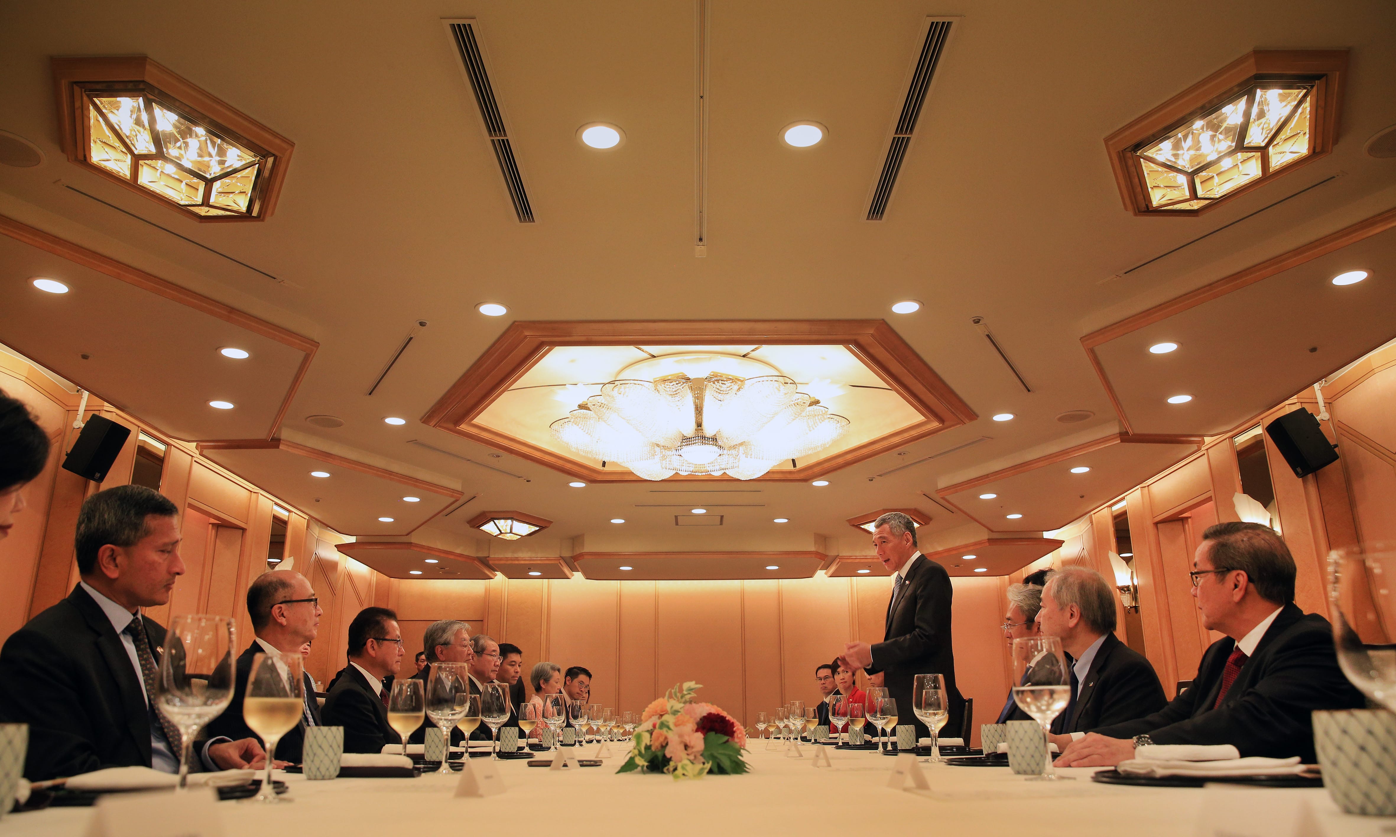 Lee Hsien Loong speaks at a long table, flanked by formally dressed attendees in a warm-toned room.