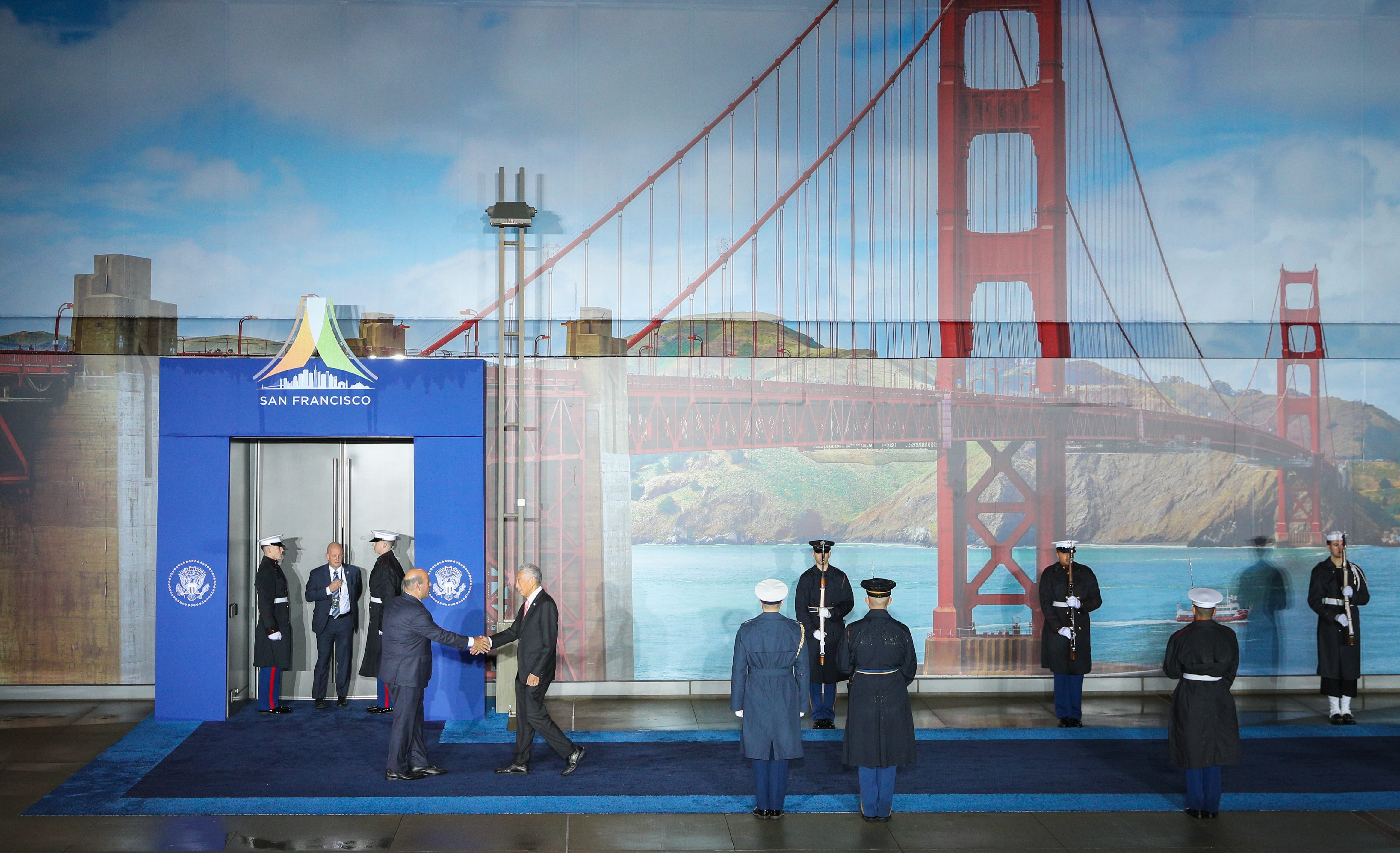 Men in suits shaking hands before guards, against Golden Gate Bridge and San Francisco backdrop.