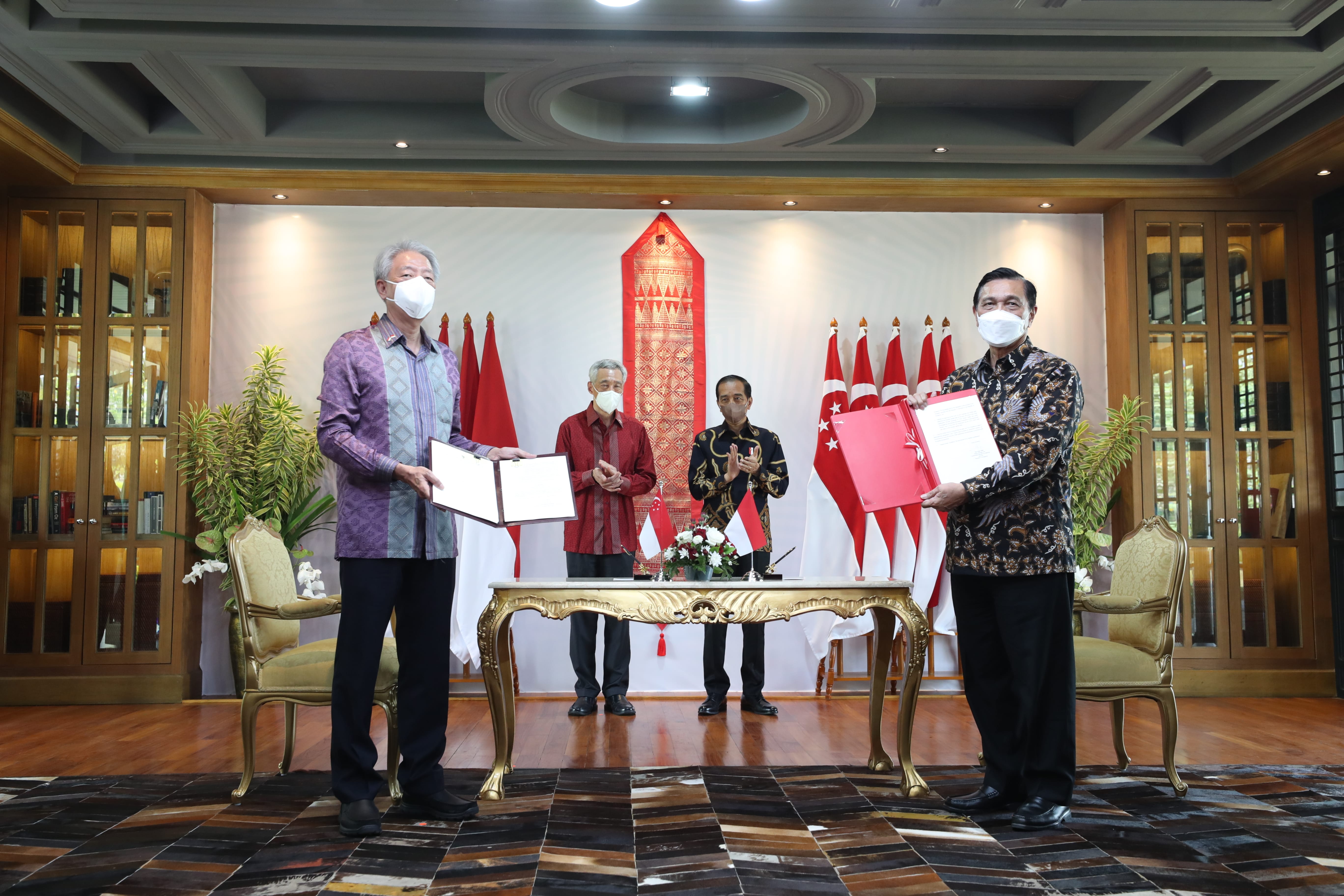 Four masked officials hold documents at a signing ceremony with flags and a table.