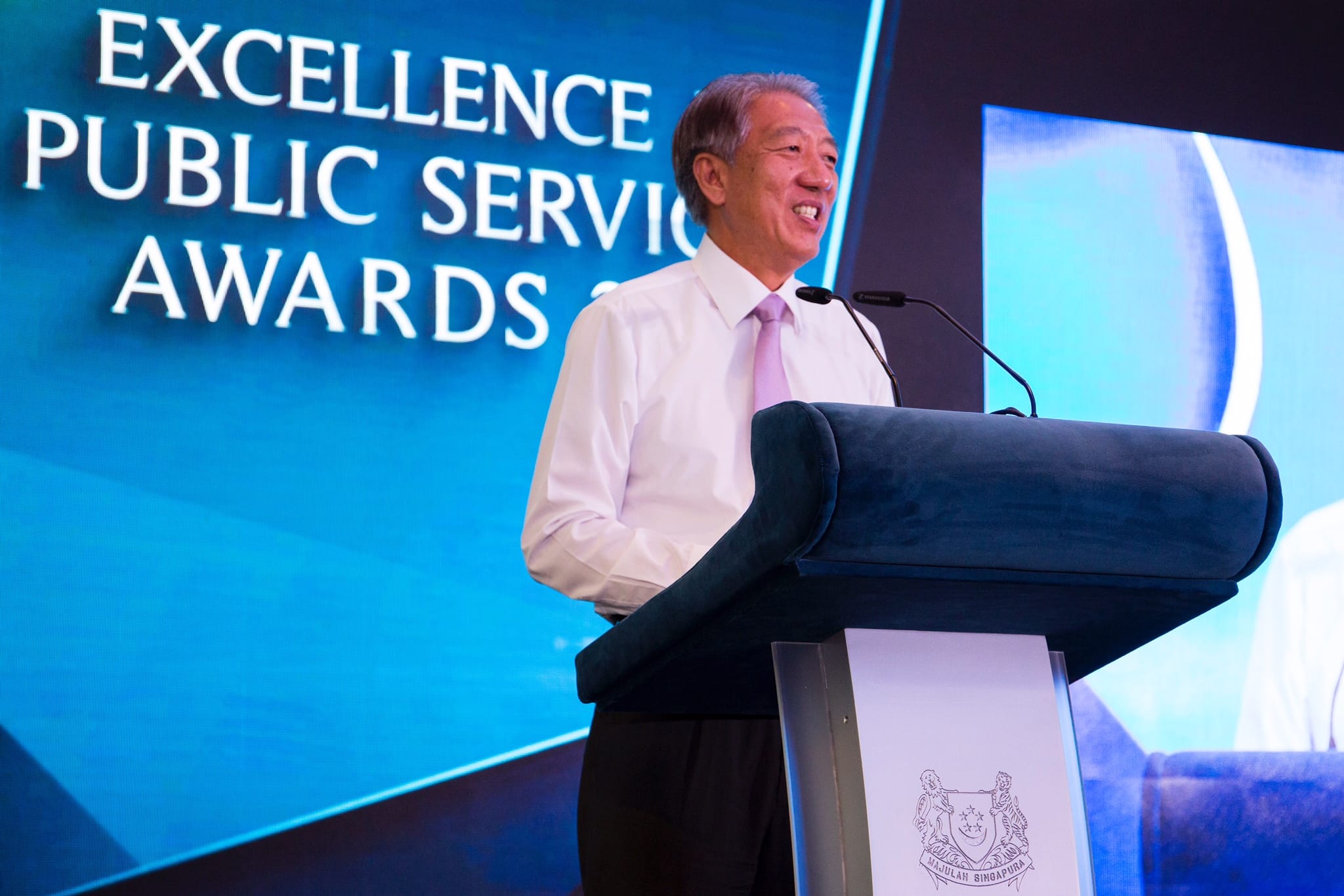 Man at podium with mic. "Excellence Public Service Awards" text and Singapore coat of arms visible.