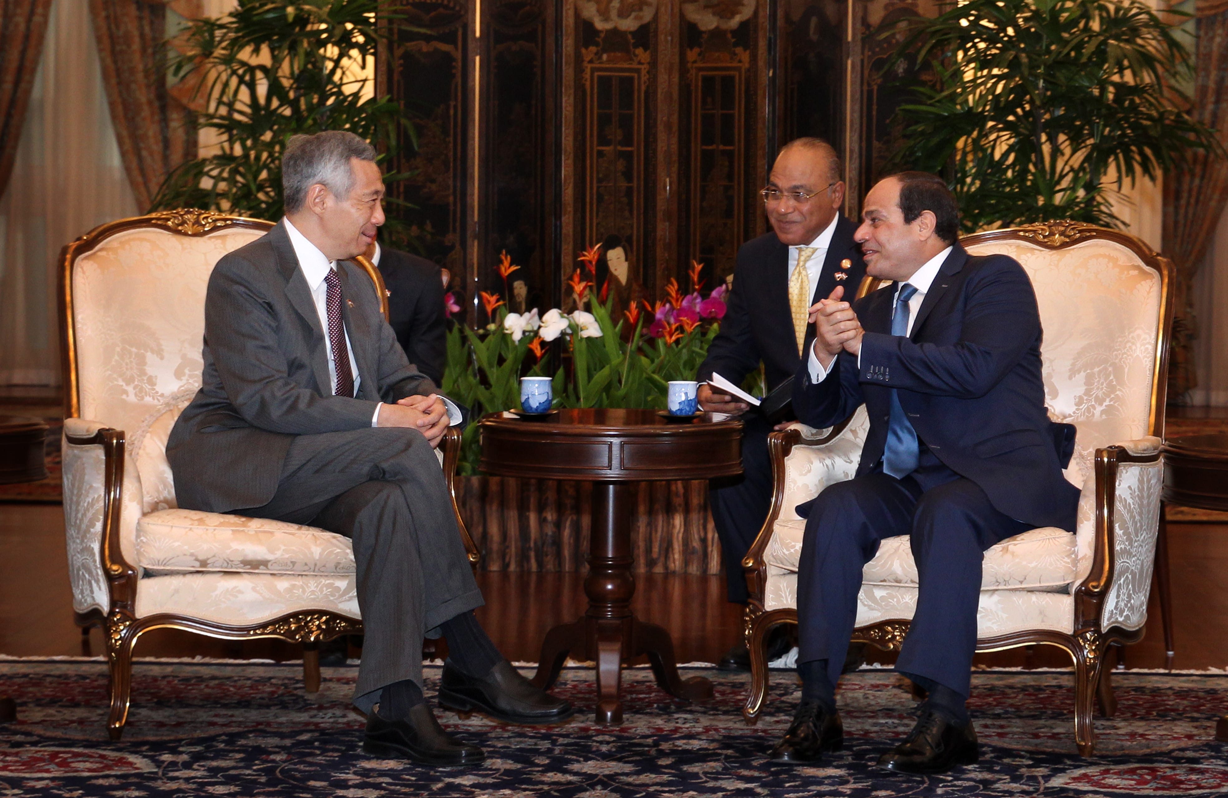 Three men in suits sit in ornate chairs around a low table with flowers.